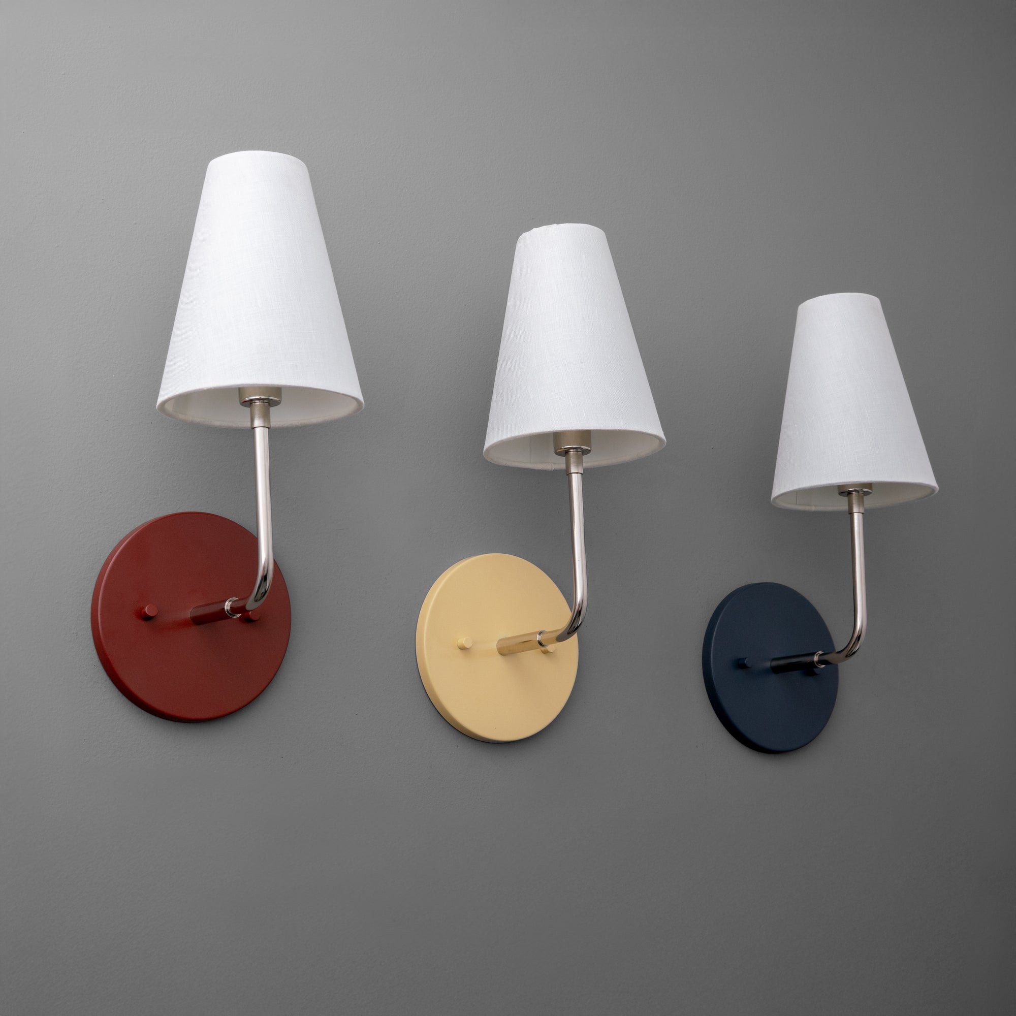 Three polished nickel wall sconces with white fabric shades, mounted on round brick red, cream, and coal blue bases against. - SCONCE MODEL NO. 3928
