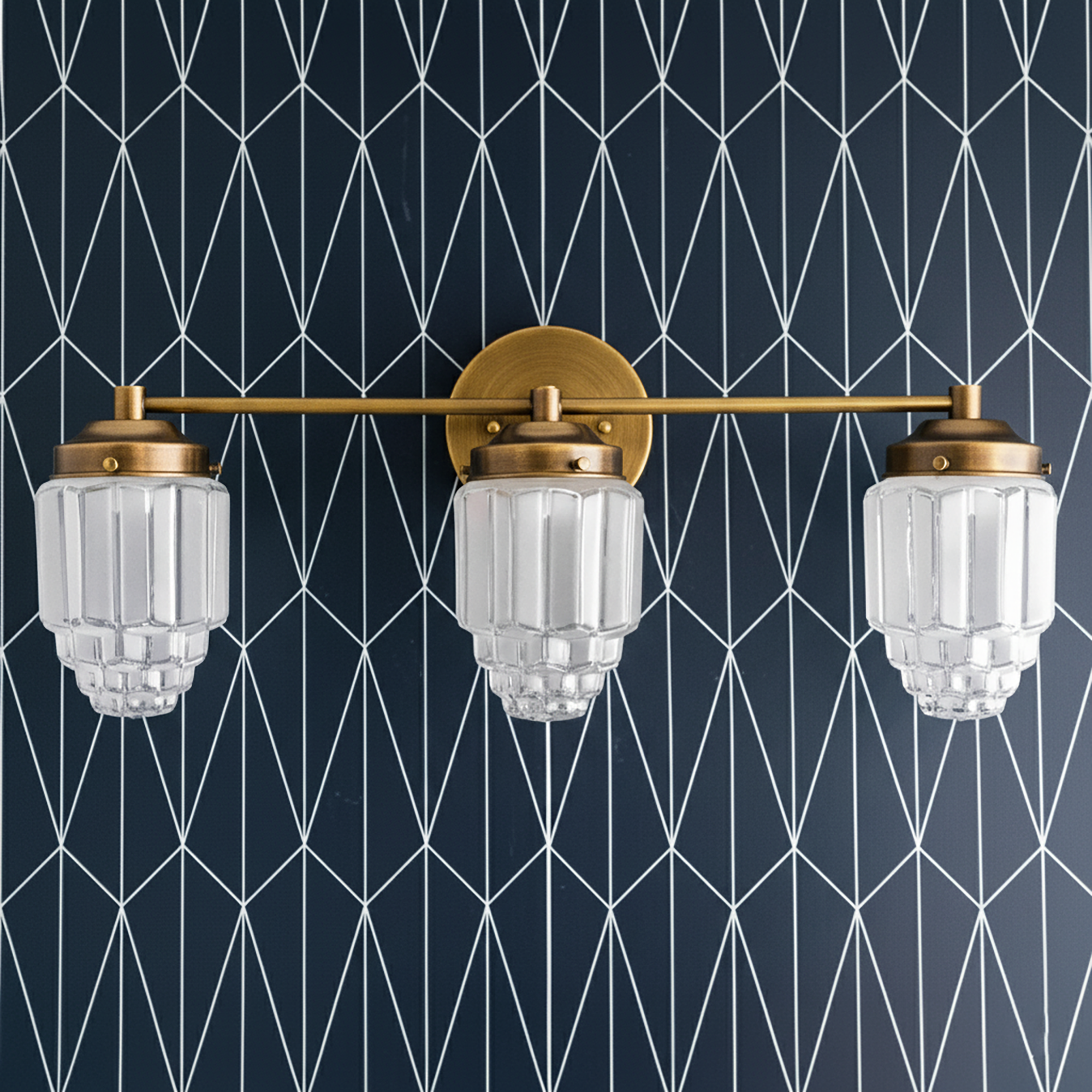 Three-bulb vanity light with raw brass finish and tiered glass globe shades against geometric wallpaper background. - VANITY LIGHT MODEL NO. 3918