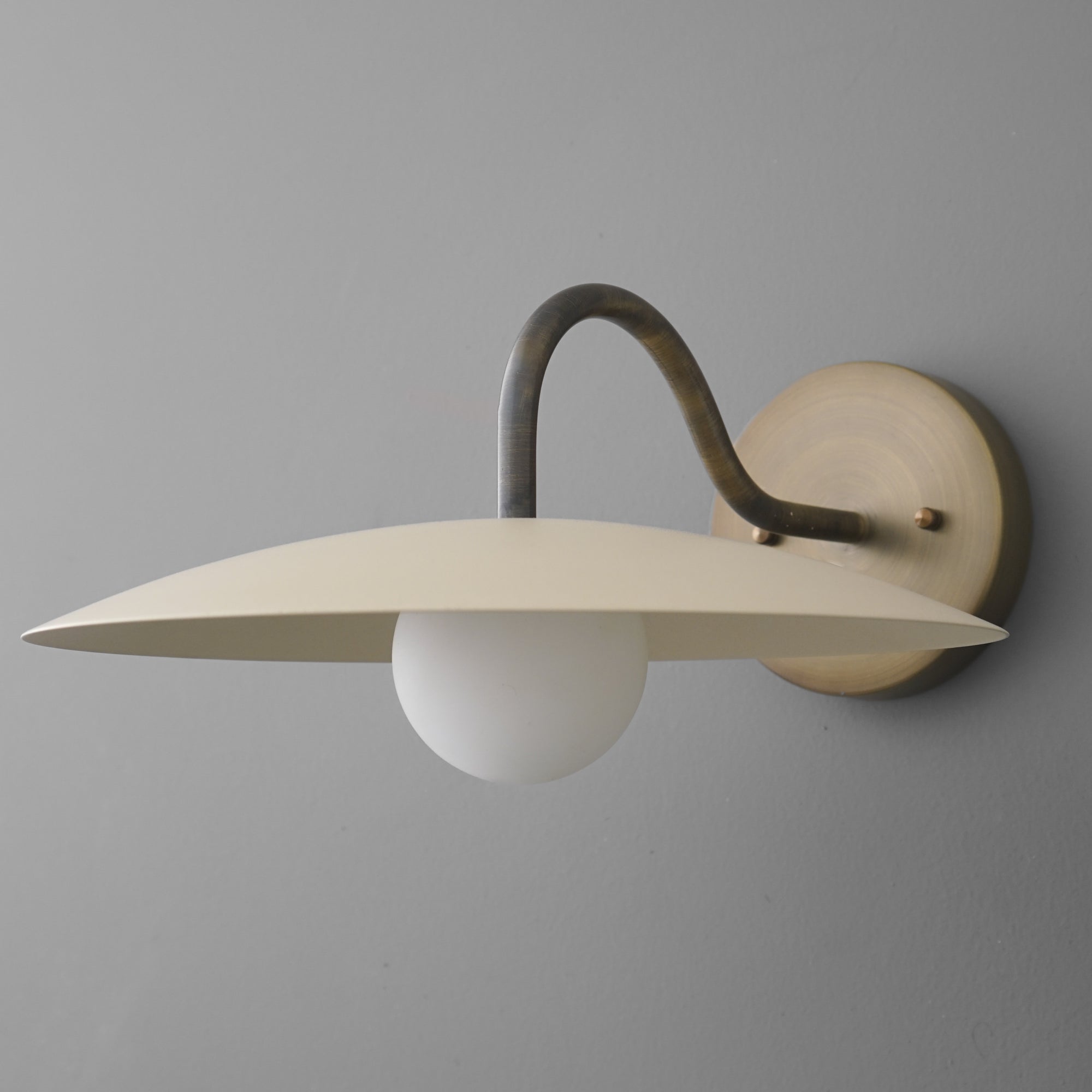 Antique brass wall sconce with curved arm, cream saucer-shaped shade, and round white glass bulb. - SCONCE MODEL NO. 2162