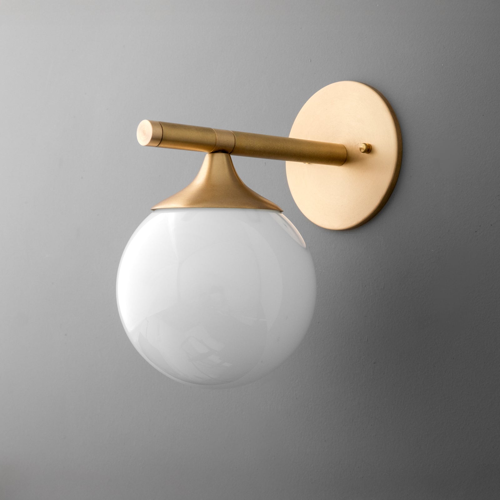 Wall sconce with raw brass finish and white frosted glass globe shade on gray wall. - SCONCE MODEL NO. 1098