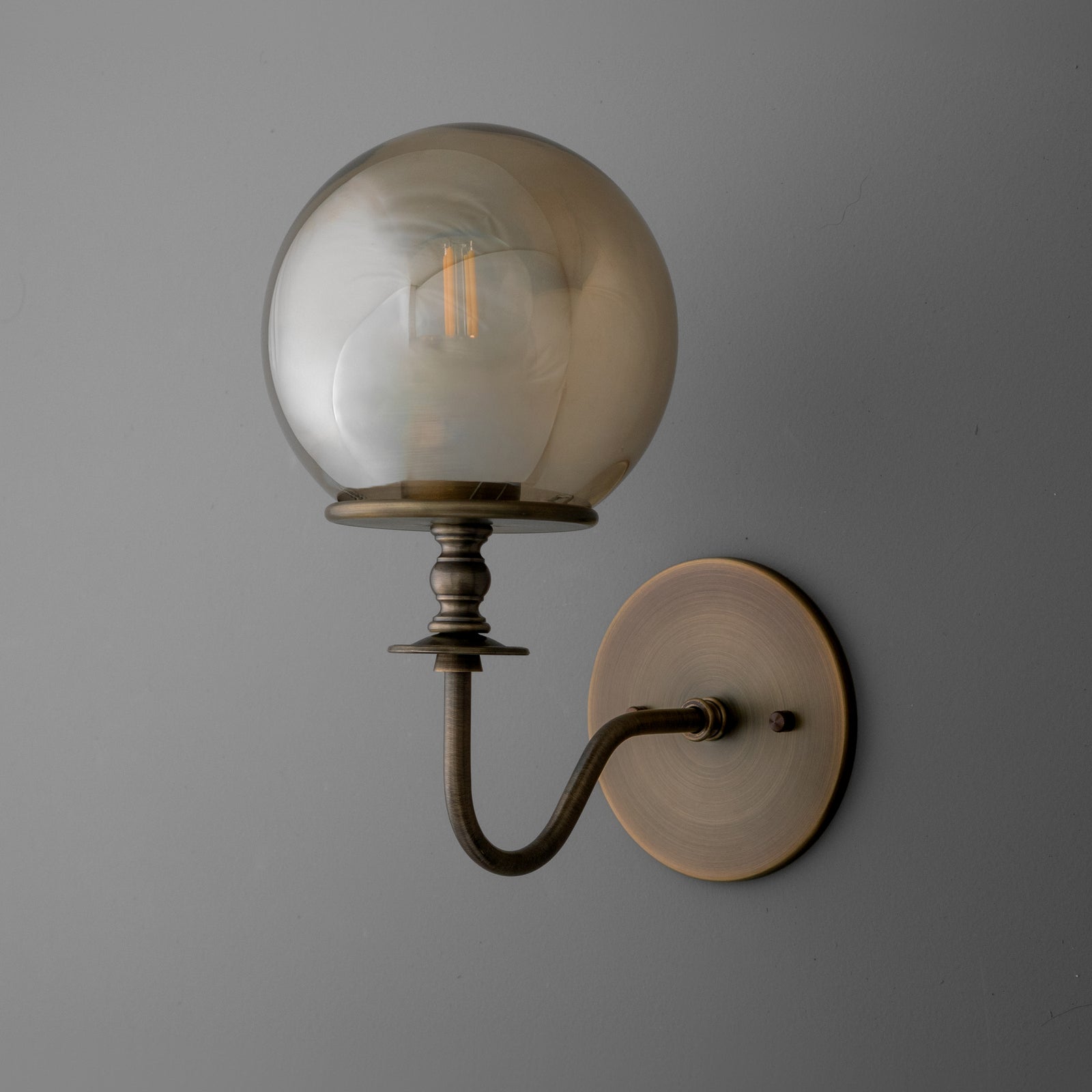Antique brass wall sconce with curved arm and smoky glass globe shade housing filament bulb. - SCONCE MODEL NO. 1029