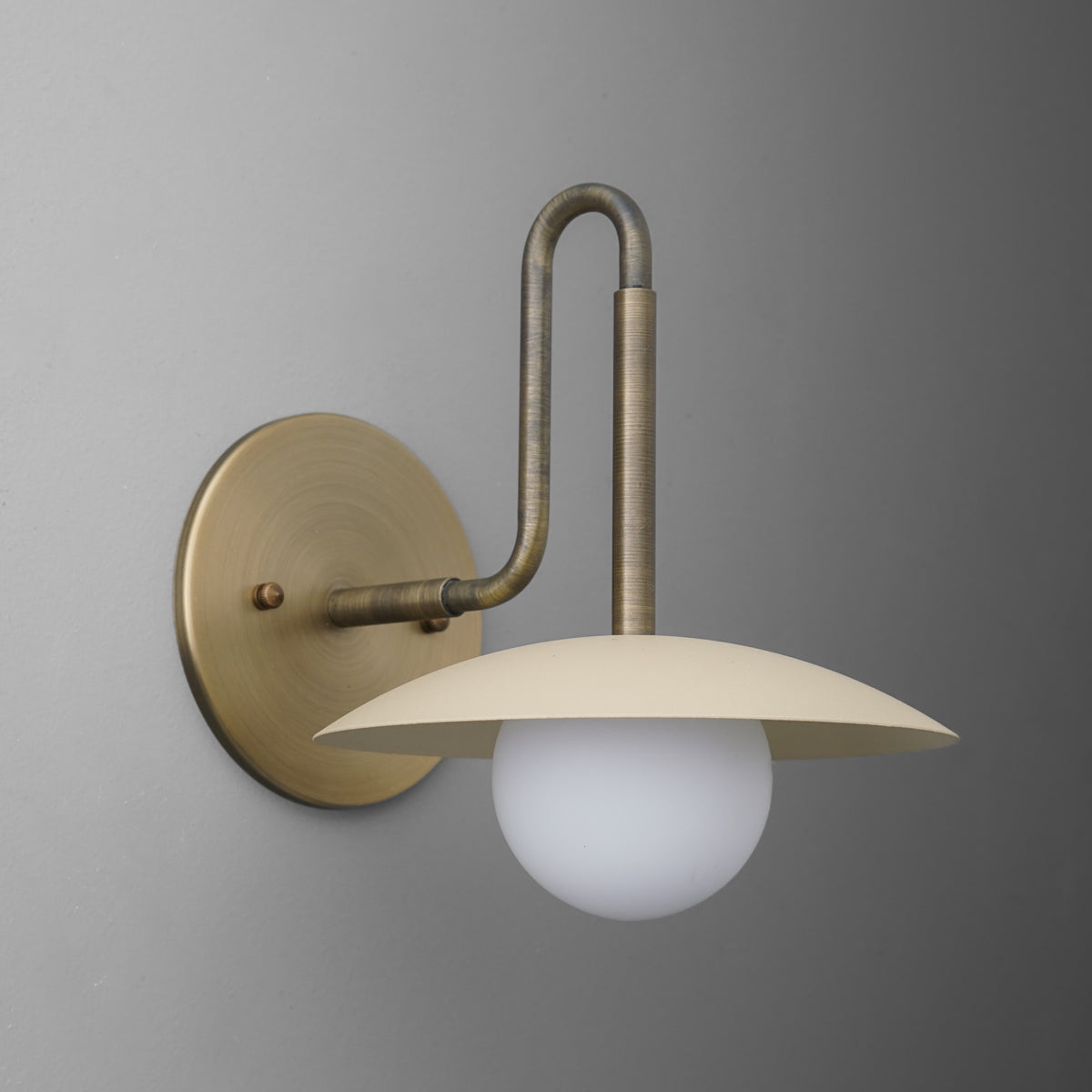Antique brass 1-bulb wall sconce with curved arm and flat, round beige shade over frosted globe bulb. - SCONCE MODEL NO. 1017