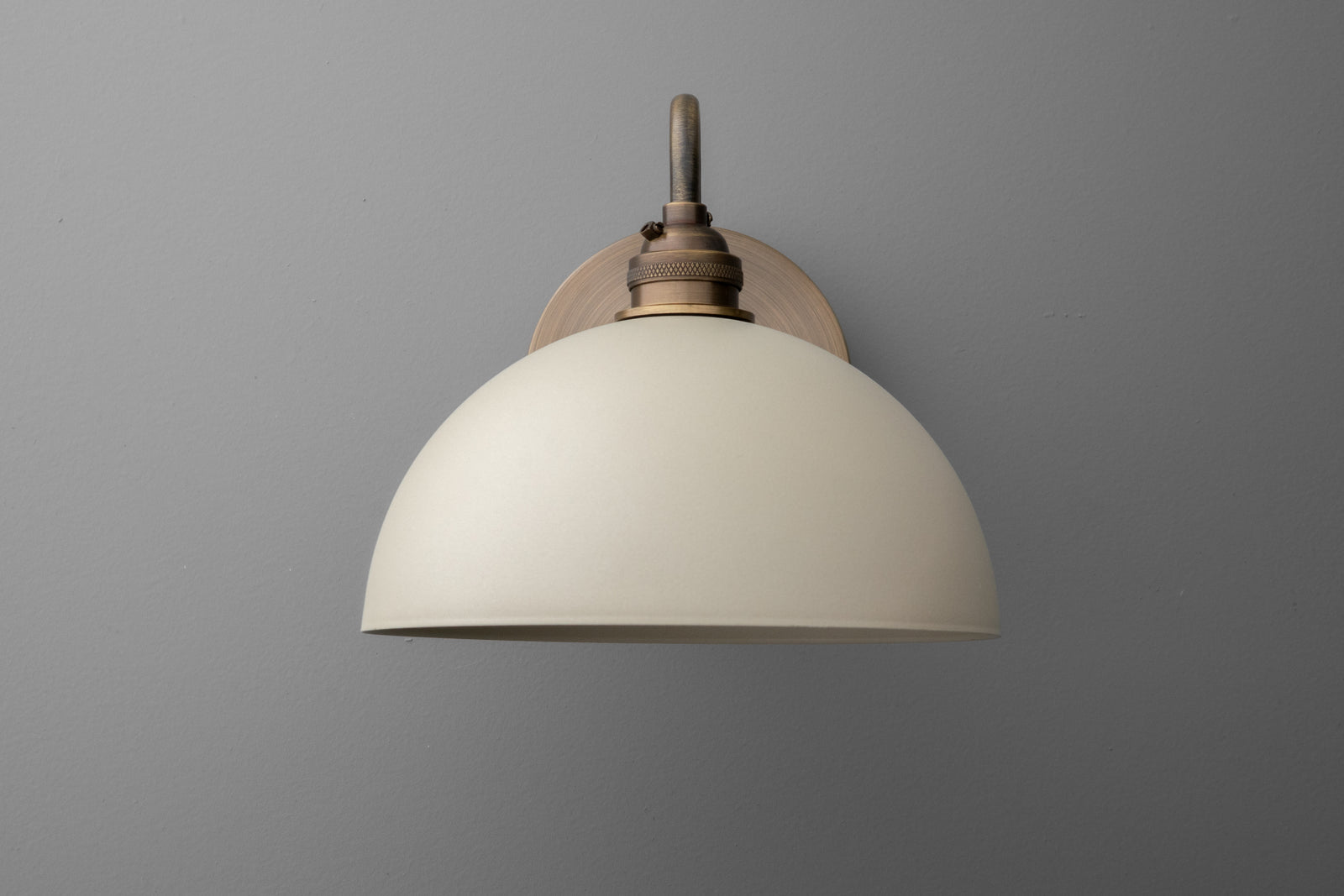 Wall sconce with cream dome shade and antique brass finish mounting on gray wall. - SCONCE MODEL NO. 9103