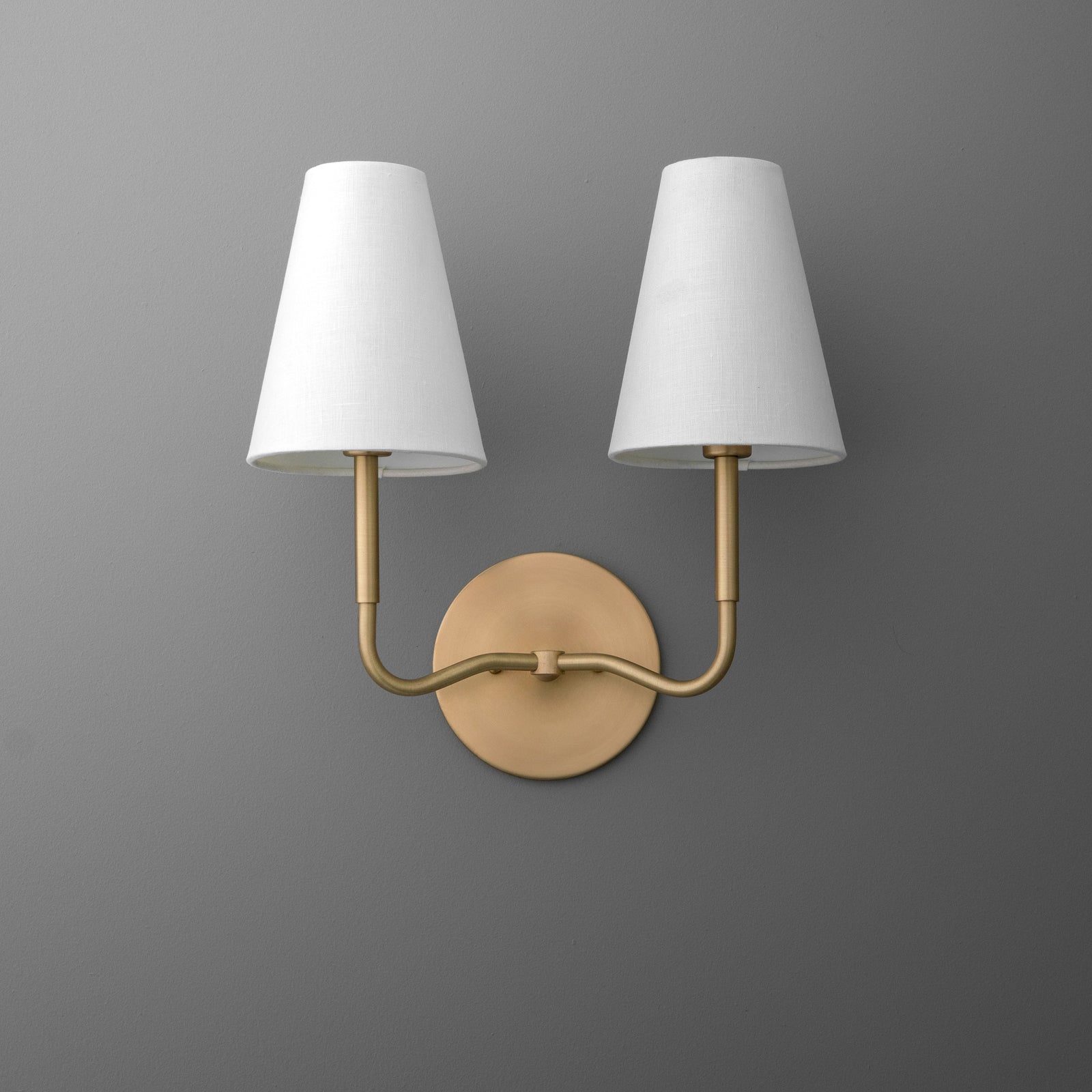 Wall-mounted sconce with two white tapered fabric shades and a curved brushed brass frame. - SCONCE MODEL NO. 9045