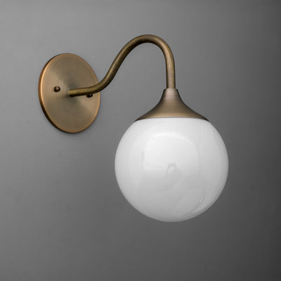 Wall-mounted sconce with antique brass curved arm and round white glass globe shade against gray wall. - SCONCE MODEL NO. 8734