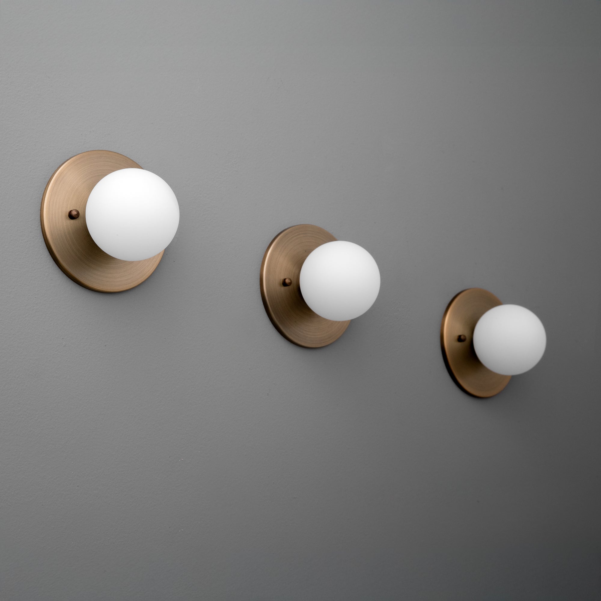 Three antique brass wall sconces with matte white 3-inch globe shades mounted on a gray wall. - Sconce Model No. 5206