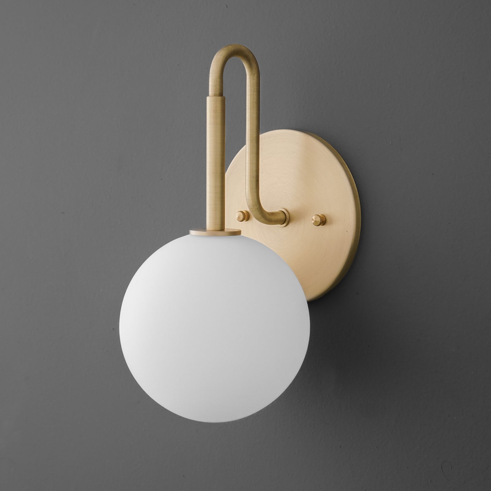 Matte white glass globe sconce with curved raw brass arm and round backplate on gray wall. - SCONCE MODEL NO. 9105