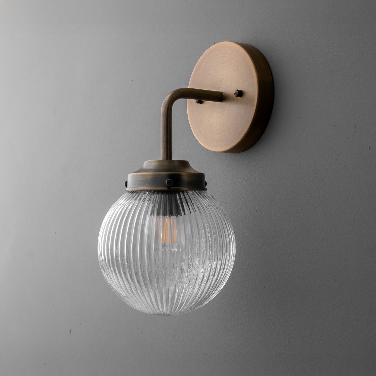 Wall sconce with antique brass finish and ribbed clear glass globe shade mounted on gray wall. - SCONCE MODEL NO. 6857
