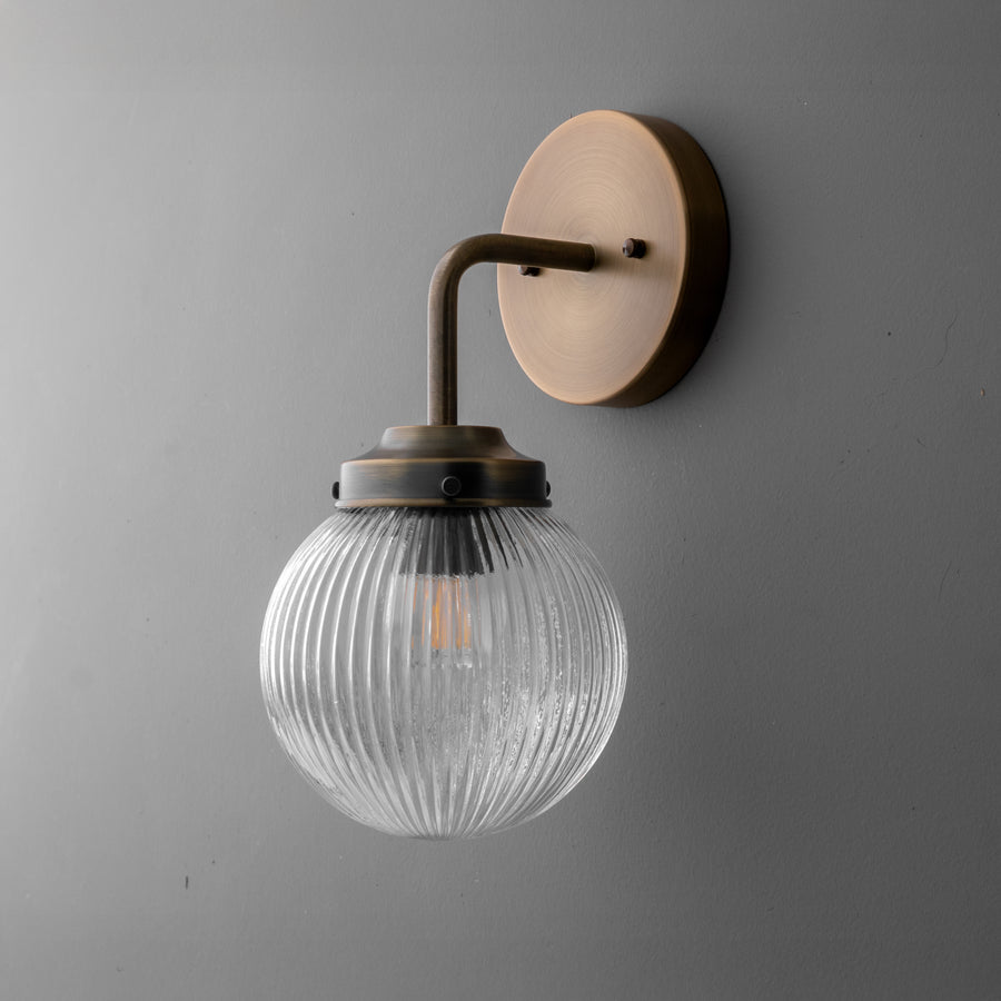 Wall sconce with antique brass finish and ribbed clear glass globe shade mounted on gray wall. - SCONCE MODEL NO. 6857