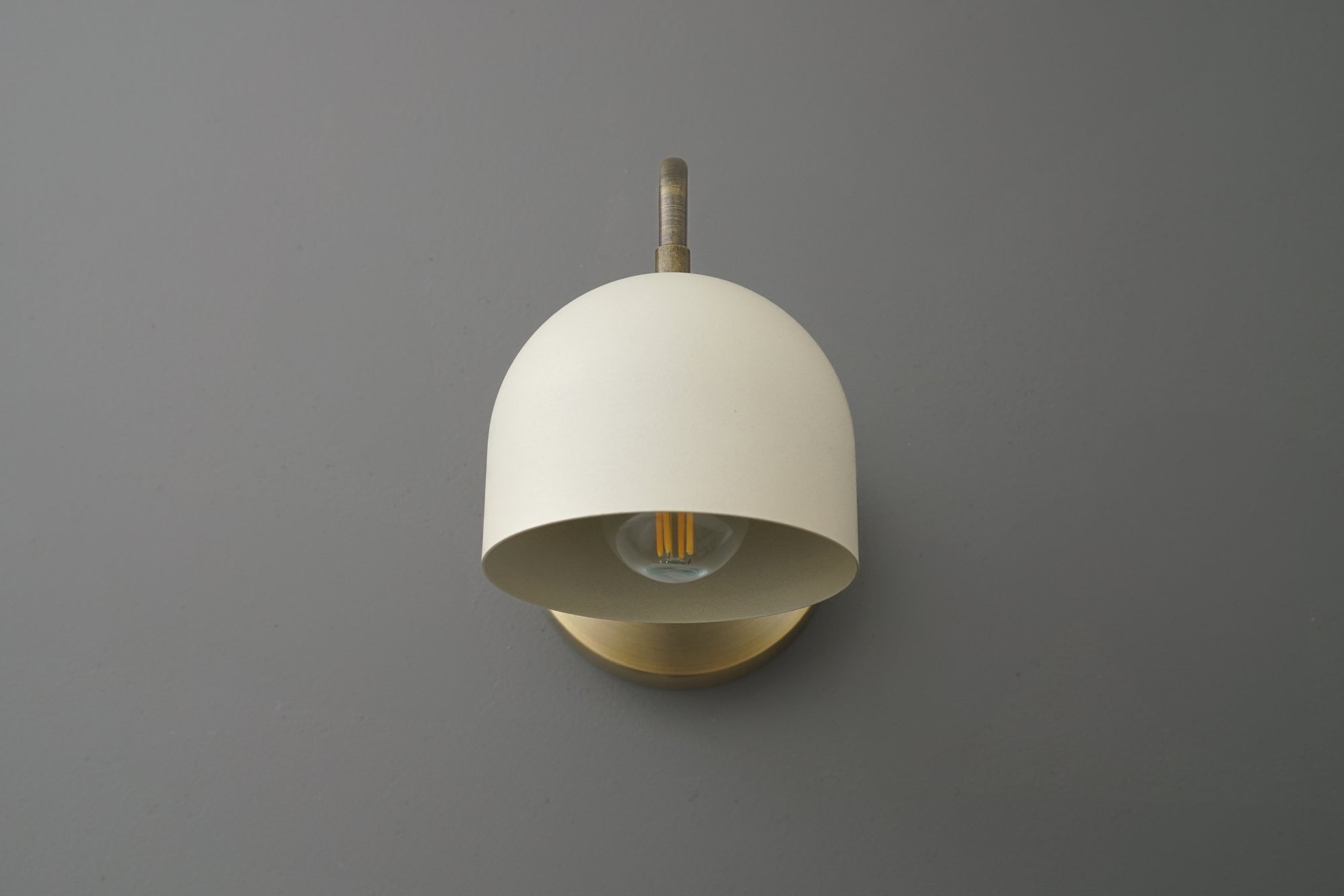 Antique brass sconce with cream half-dome shade and visible Edison bulb on gray wall. - SCONCE MODEL NO. 3628