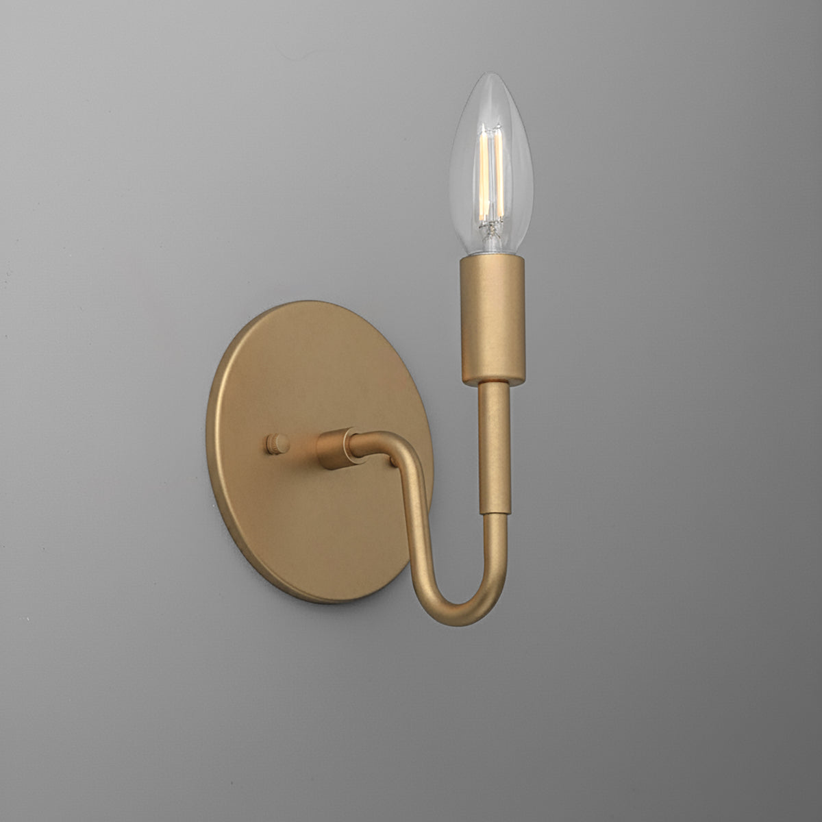 Matte gold metal wall sconce with a round base and a single curved arm holding a clear candle-shaped bulb. - SCONCE MODEL NO. 1100