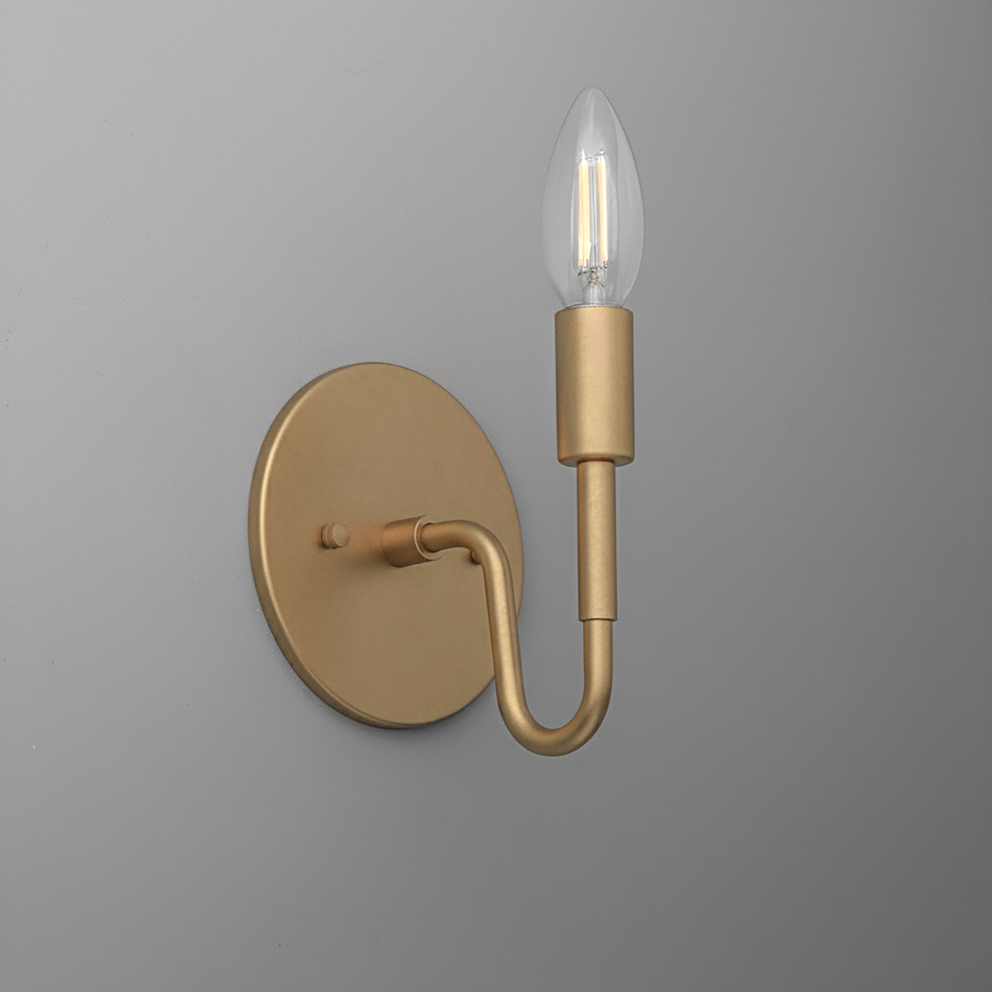 Matte gold metal wall sconce with a round base and a single curved arm holding a clear candle-shaped bulb. - SCONCE MODEL NO. 1100
