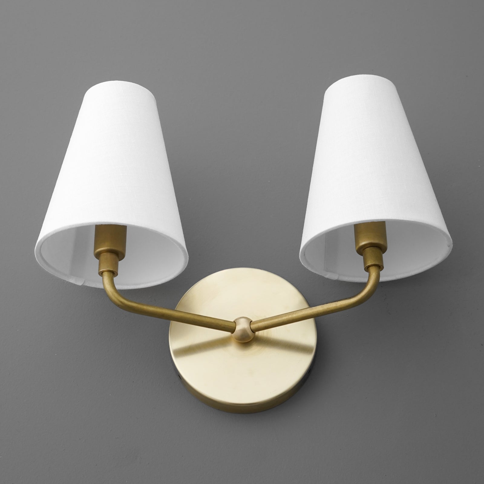 Two-bulb wall sconce with brushed brass arms and white tapered fabric shades on gray background. - SCONCE MODEL NO. 0823