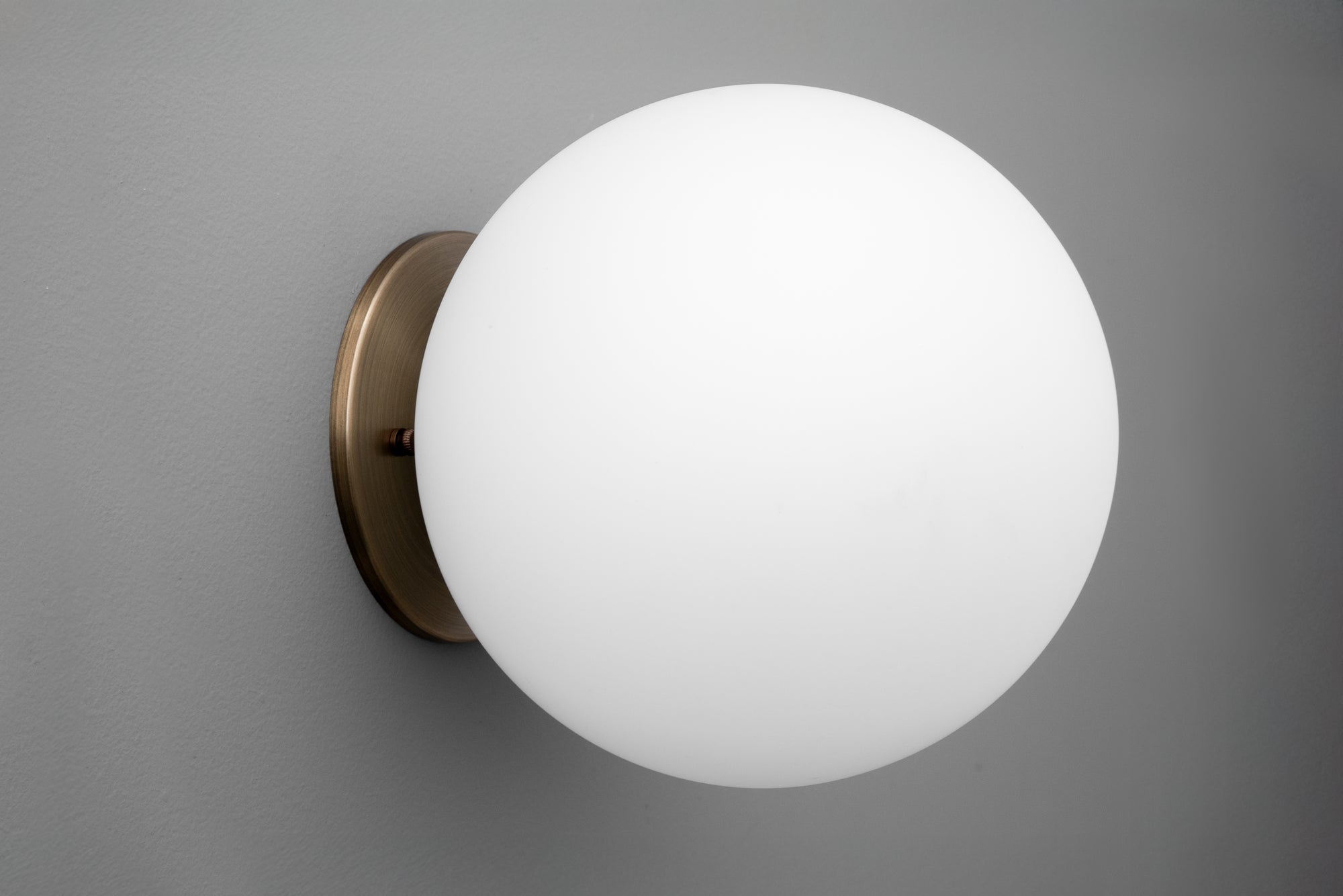 Minimalist wall sconce with a matte white glass globe and antique brass mount on gray wall. - Sconce Model No. 5206