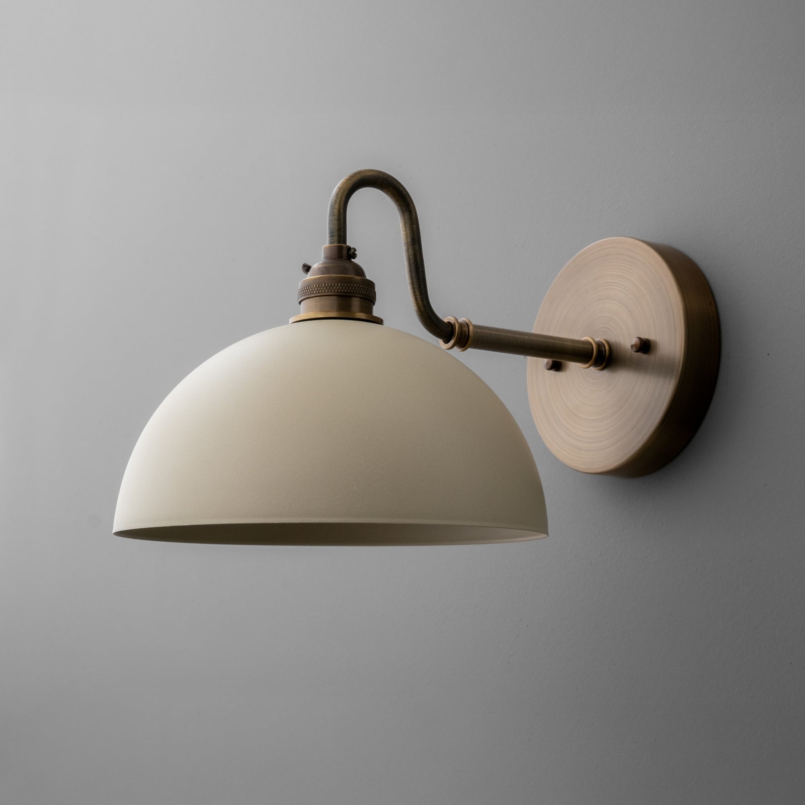 Antique brass wall sconce with curved arm and cream dome shade against gray wall. - SCONCE MODEL NO. 9103