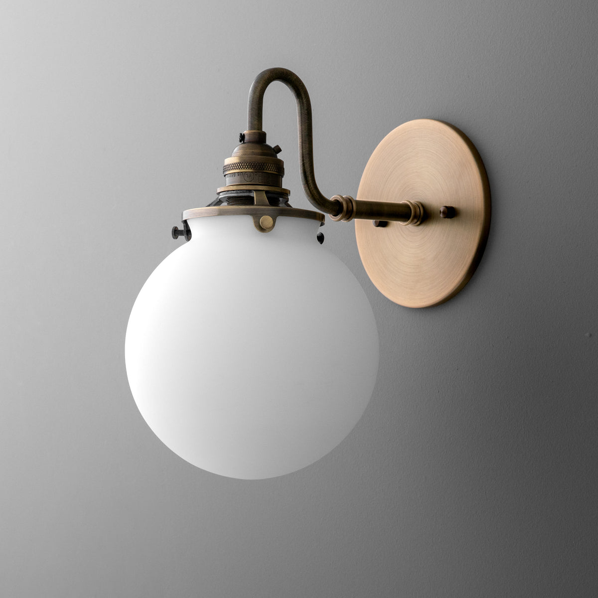 Single-light sconce in Antique Brass with a hooked arm and exposed socket holding a round opal glass globe, on a round wall plate. - SCONCE MODEL NO. 5994
