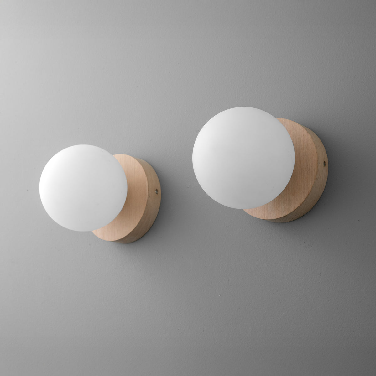 Two wall sconces with round matte white glass globes and light beechwood bases mounted on a light gray wall. - SCONCE MODEL NO. 4943