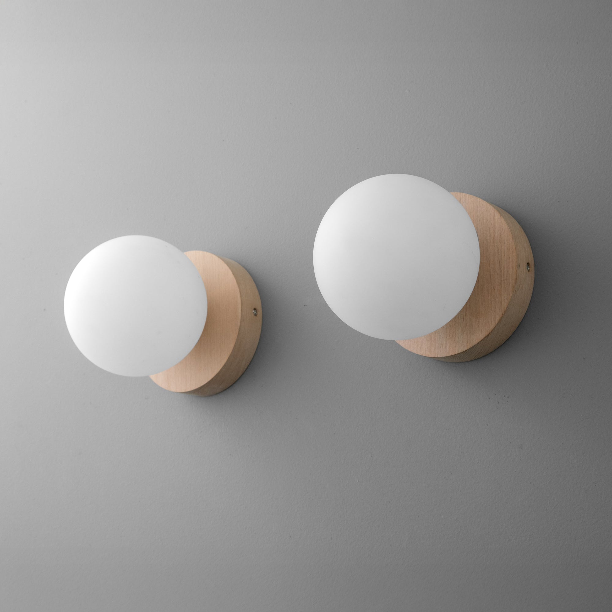 Two wall sconces with round matte white glass globes and light beechwood bases mounted on a light gray wall. - SCONCE MODEL NO. 4943