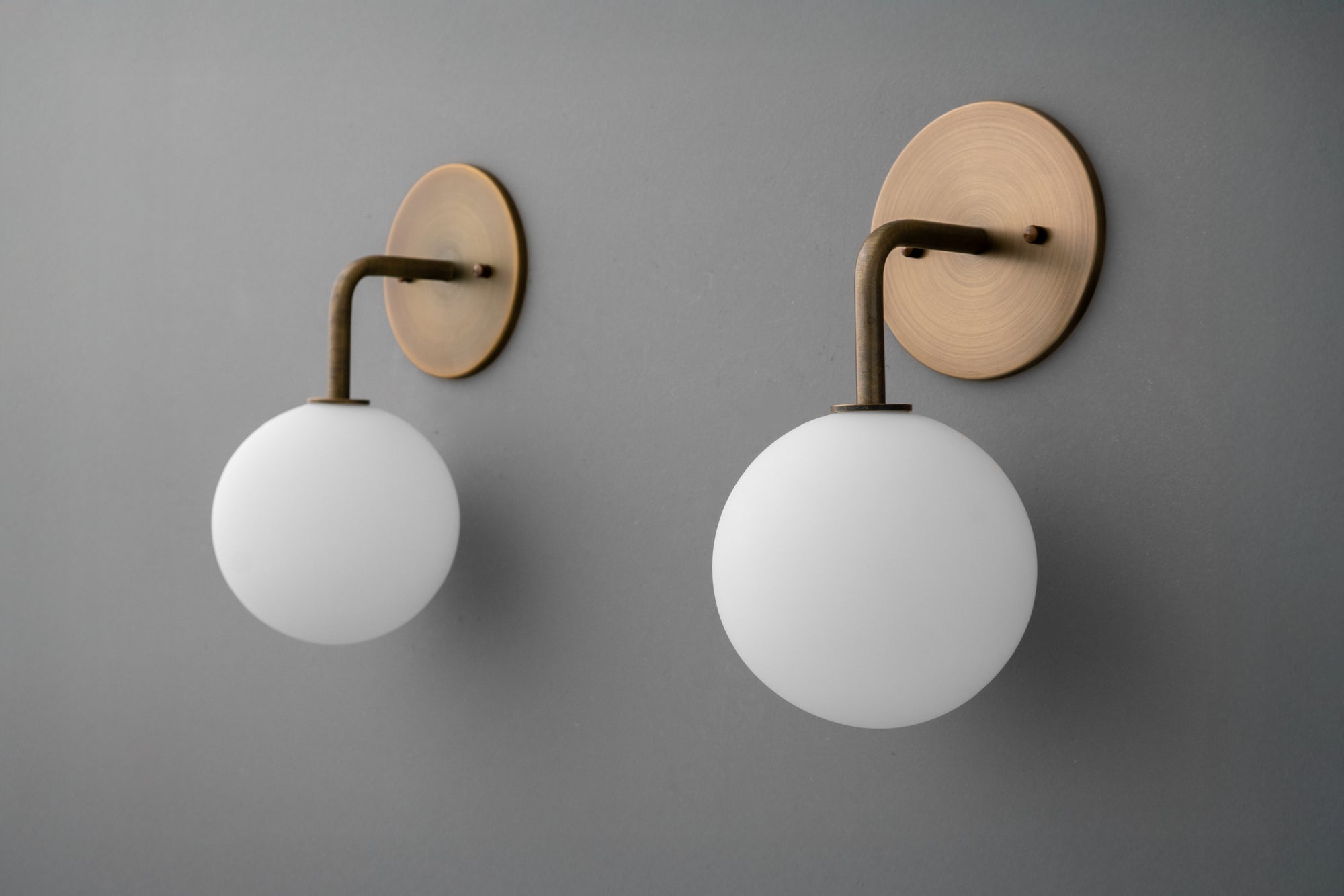 Pair of wall sconces with round antique brass mounts and curved arms, featuring large white opal glass globes. - SCONCE MODEL NO. 7660