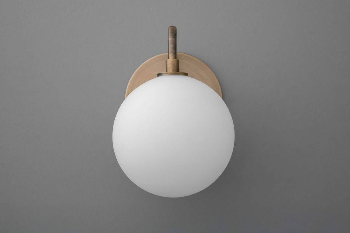 Minimalist wall sconce with frosted white glass globe and antique brass mount on gray wall. - SCONCE MODEL NO. 8637
