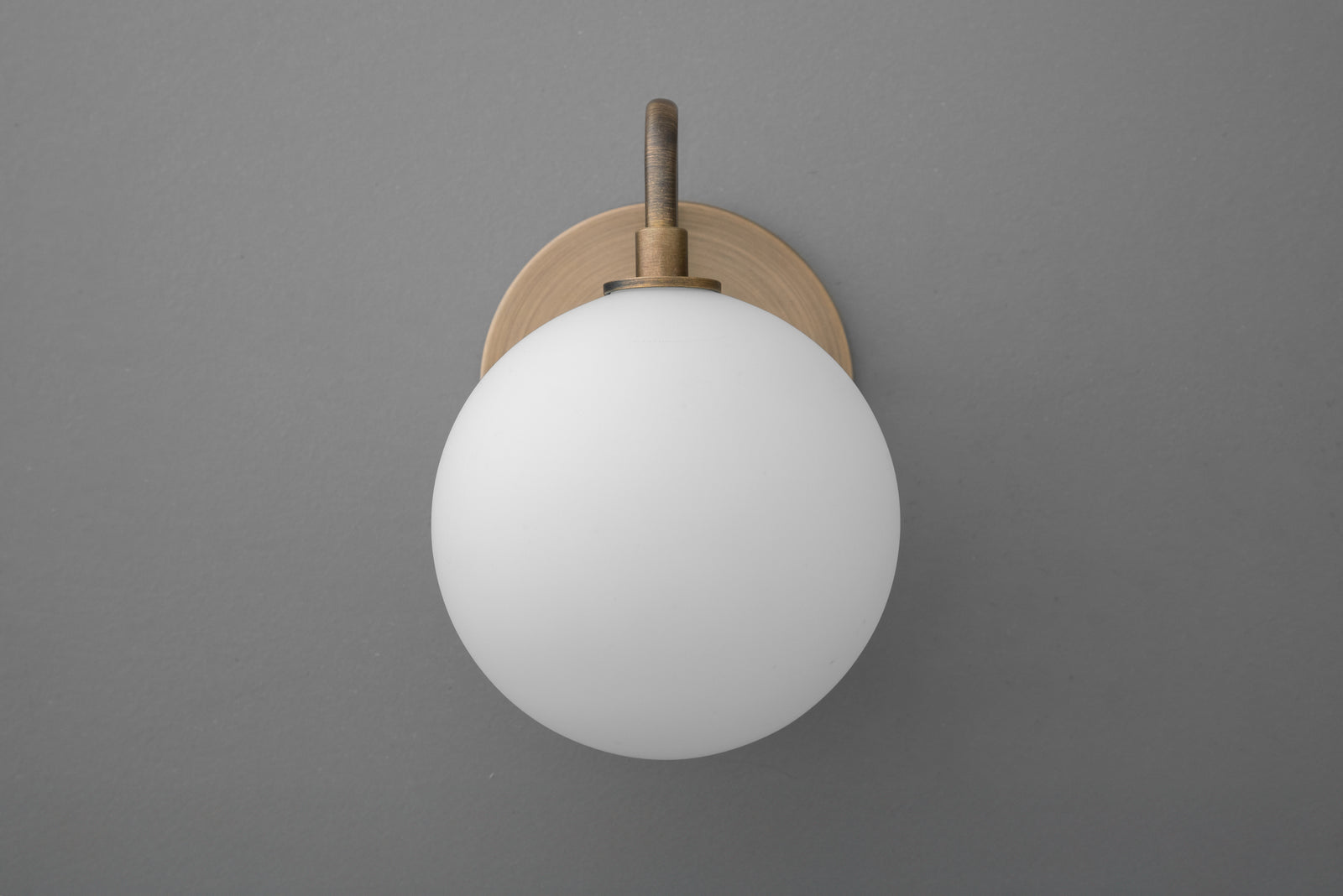 Minimalist wall sconce with frosted white glass globe and antique brass mount on gray wall. - SCONCE MODEL NO. 8637