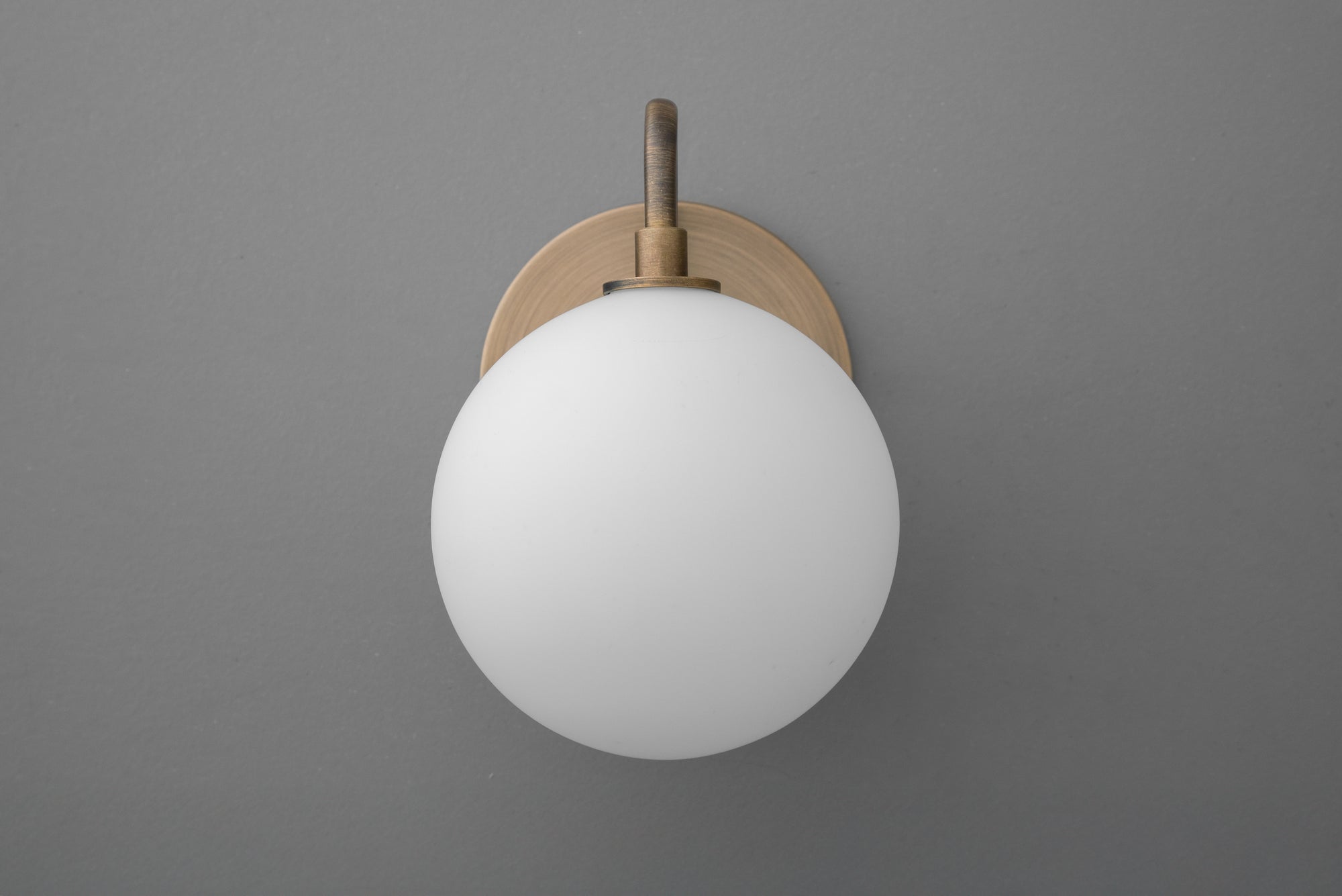 Minimalist wall sconce with frosted white glass globe and antique brass mount on gray wall. - SCONCE MODEL NO. 8637