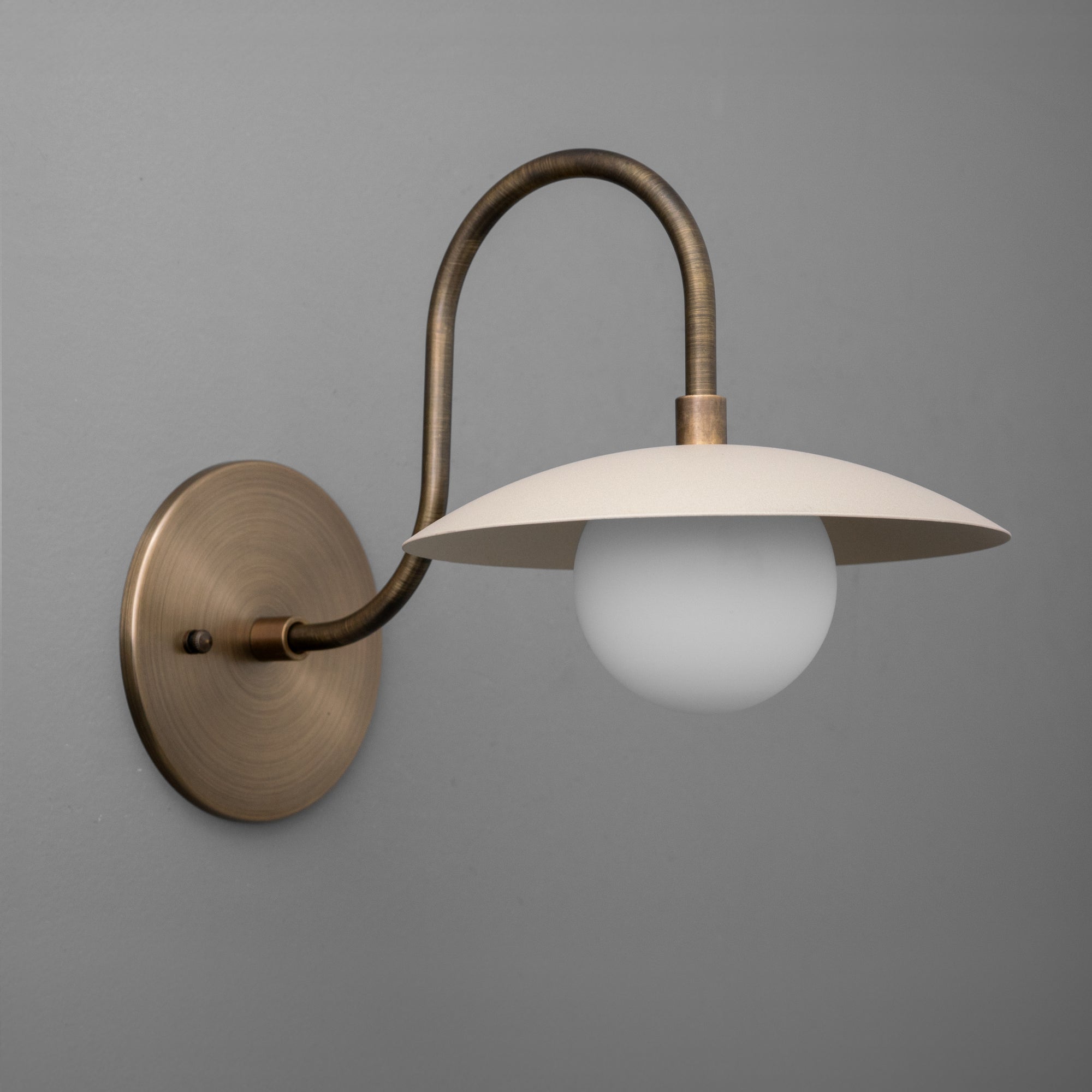 Wall sconce with antique brass curved arm, cream metal shade, and frosted globe bulb. - SCONCE MODEL NO. 3799