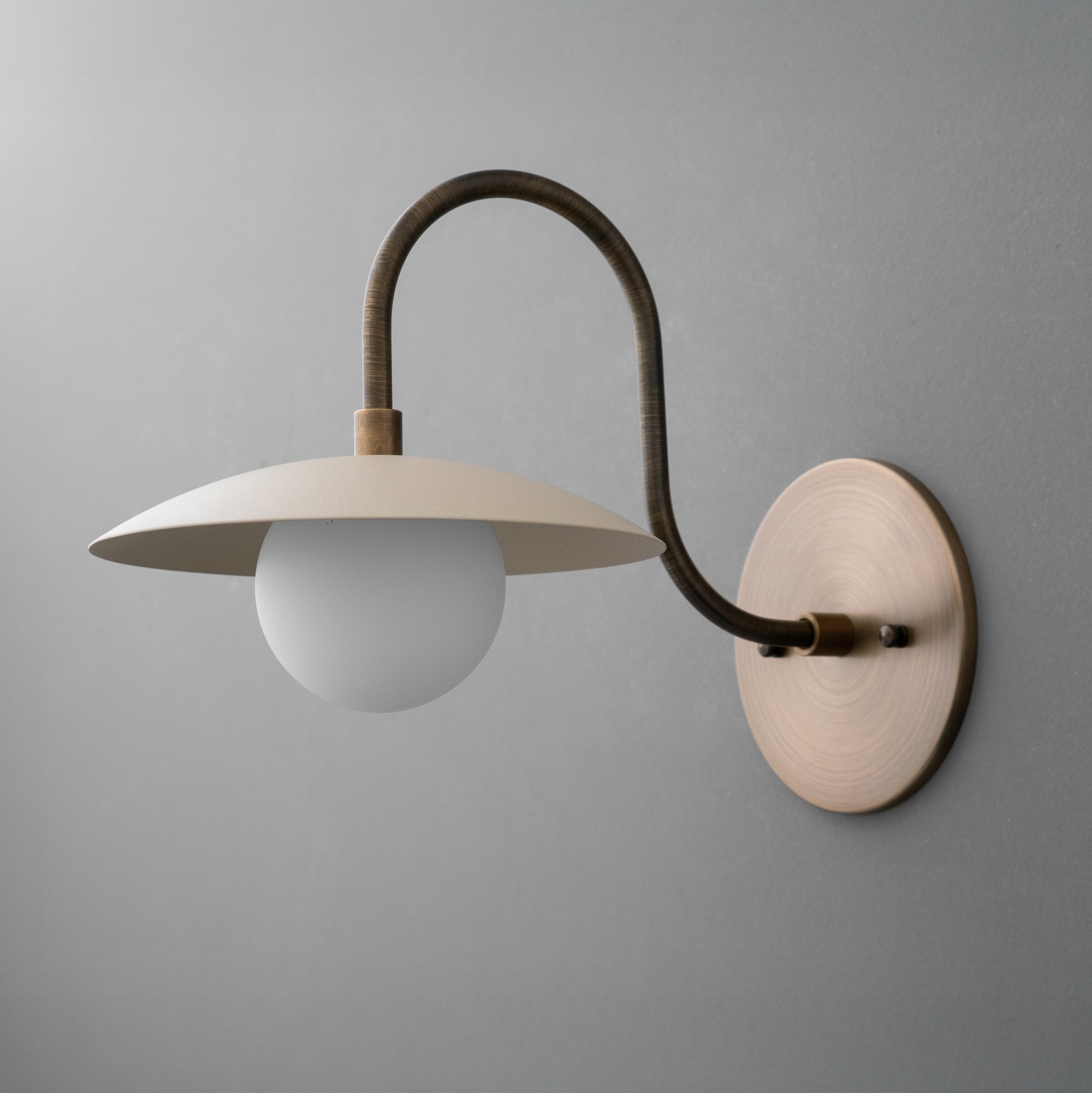 Wall-mounted sconce with a cream-colored conical shade, frosted globe bulb, and curved antique brass arm. - SCONCE MODEL NO. 3799