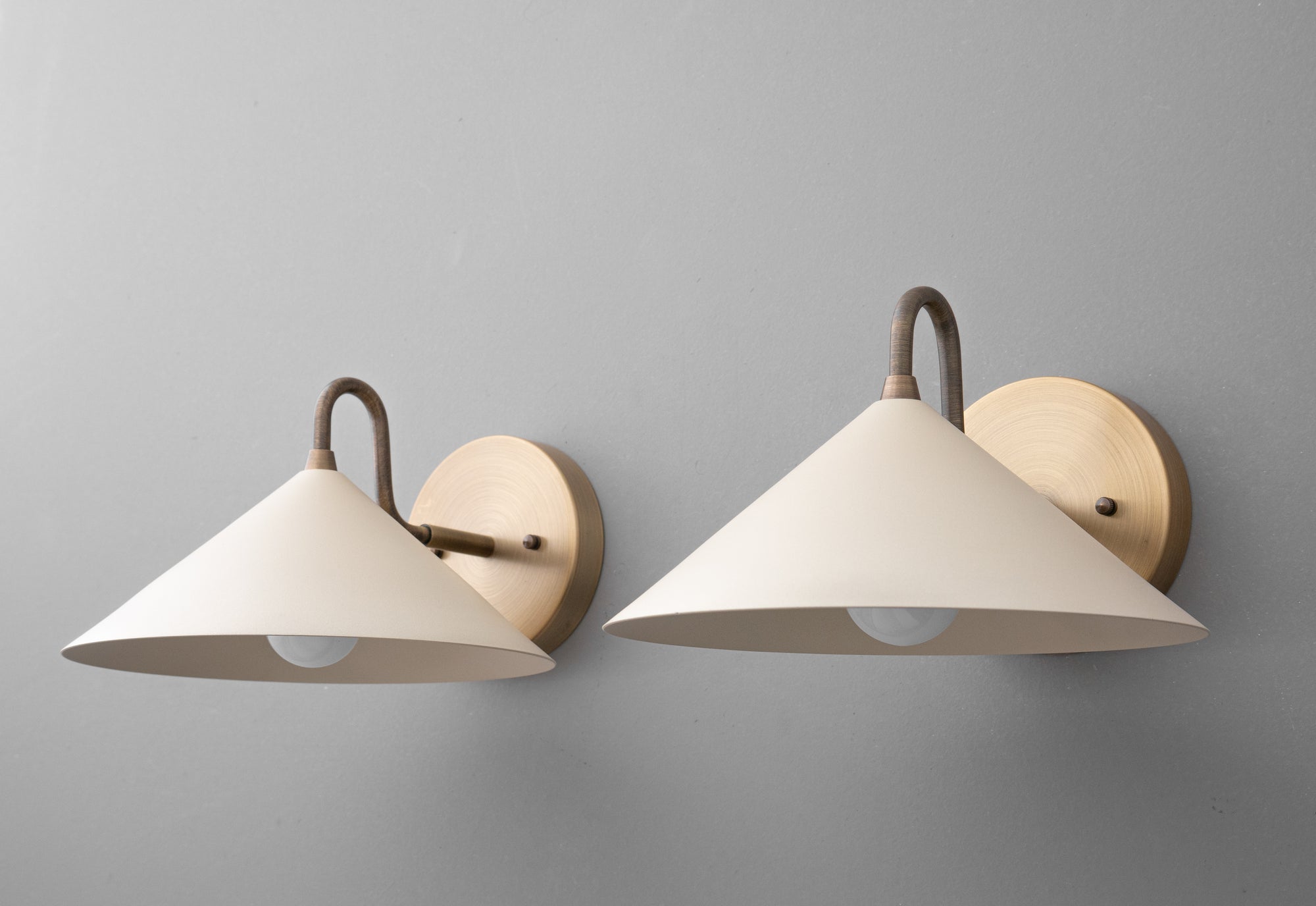 Two wall sconces with antique brass mounts and conical cream fabric shades against a gray wall background. - SCONCE MODEL NO. 6528