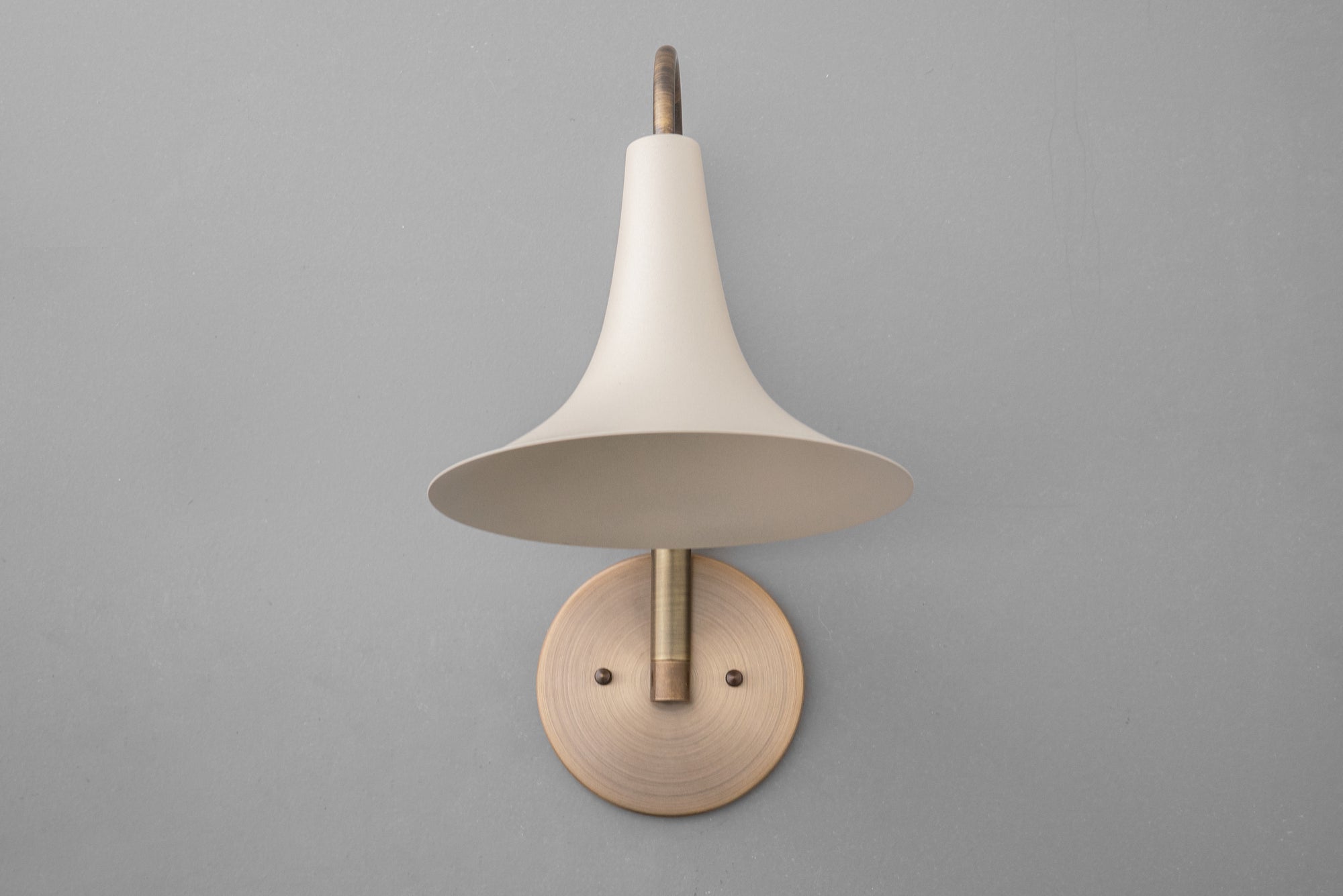 Antique brass wall sconce with a cream flared metal shade and round mounting plate on gray wall. - SCONCE MODEL NO. 3851