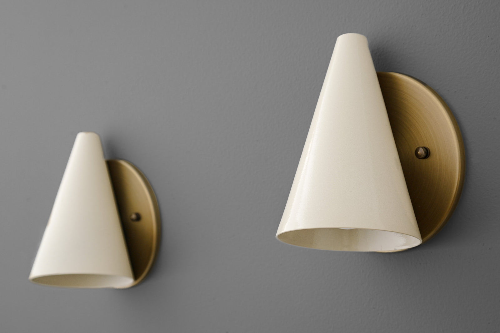 Antique brass wall sconce with a cream tapered conical shade mounted on a round backplate against a gray wall. - SCONCE MODEL NO. 5950