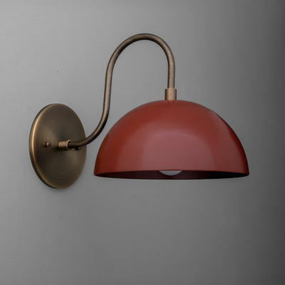 Antique brass wall sconce with curved arm and dome-shaped brick red shade mounted on gray wall. - SCONCE MODEL NO. 9092