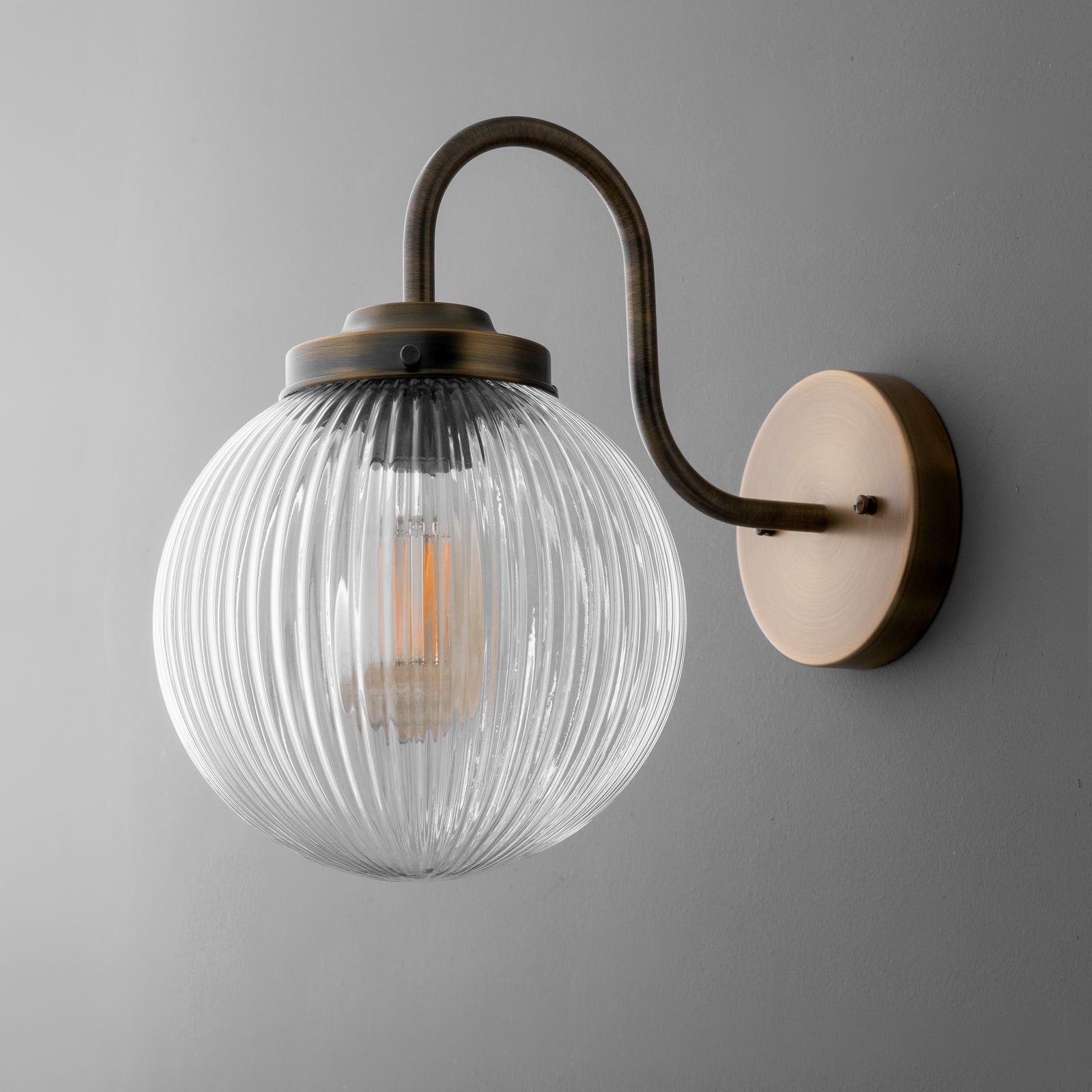 Wall sconce with ribbed clear glass globe shade and curved antique brass arm mounted on round beige base. - SCONCE MODEL NO. 1009