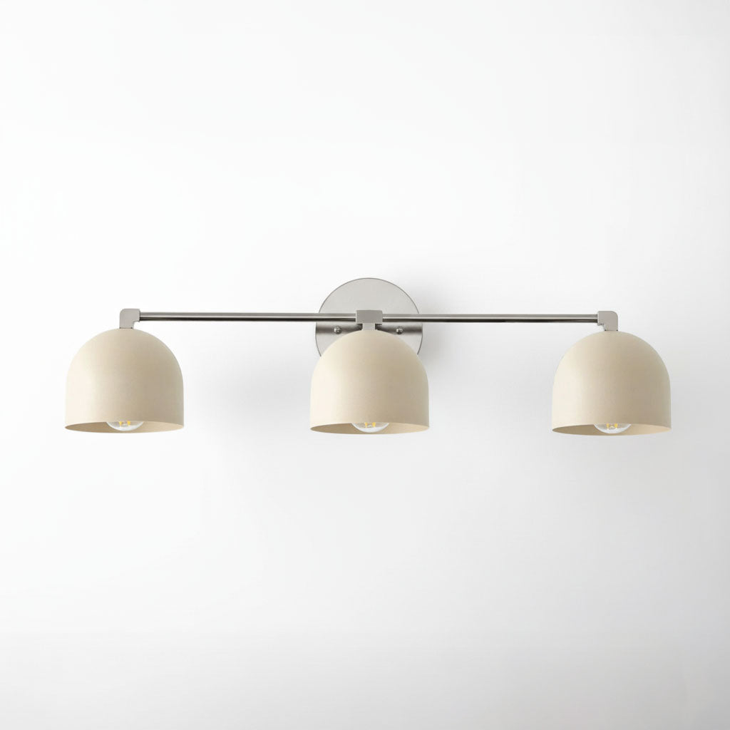 Brushed nickel vanity light with three cream dome shades mounted on a horizontal bar. - VANITY LIGHT MODEL NO. 7565