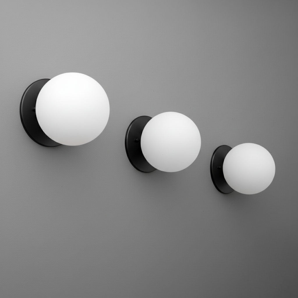 Three black metal wall sconces with frosted white glass globes in small, medium, and large sizes mounted on a gray wall. - Sconce Model No. 5206