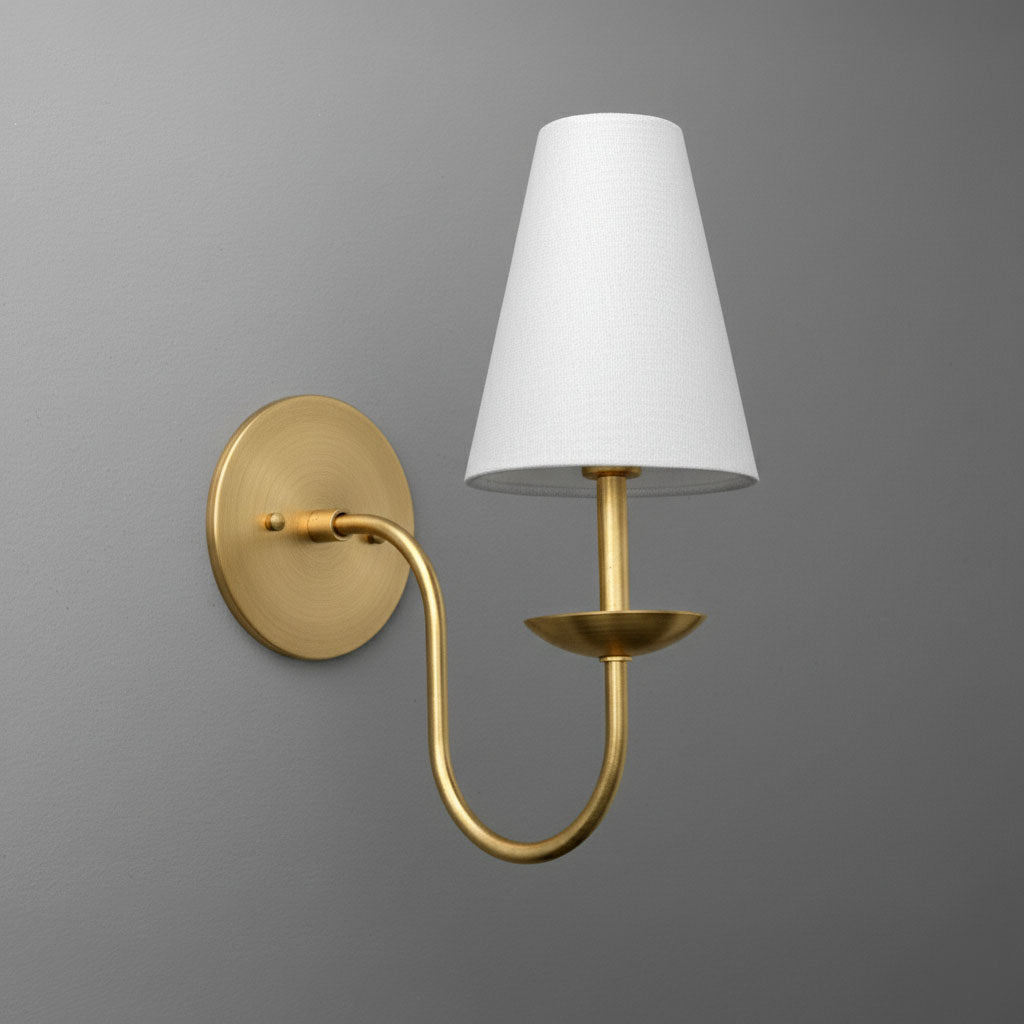 Antique brass wall sconce with curved arm and white tapered fabric shade mounted on gray wall. - SCONCE MODEL NO. 9867