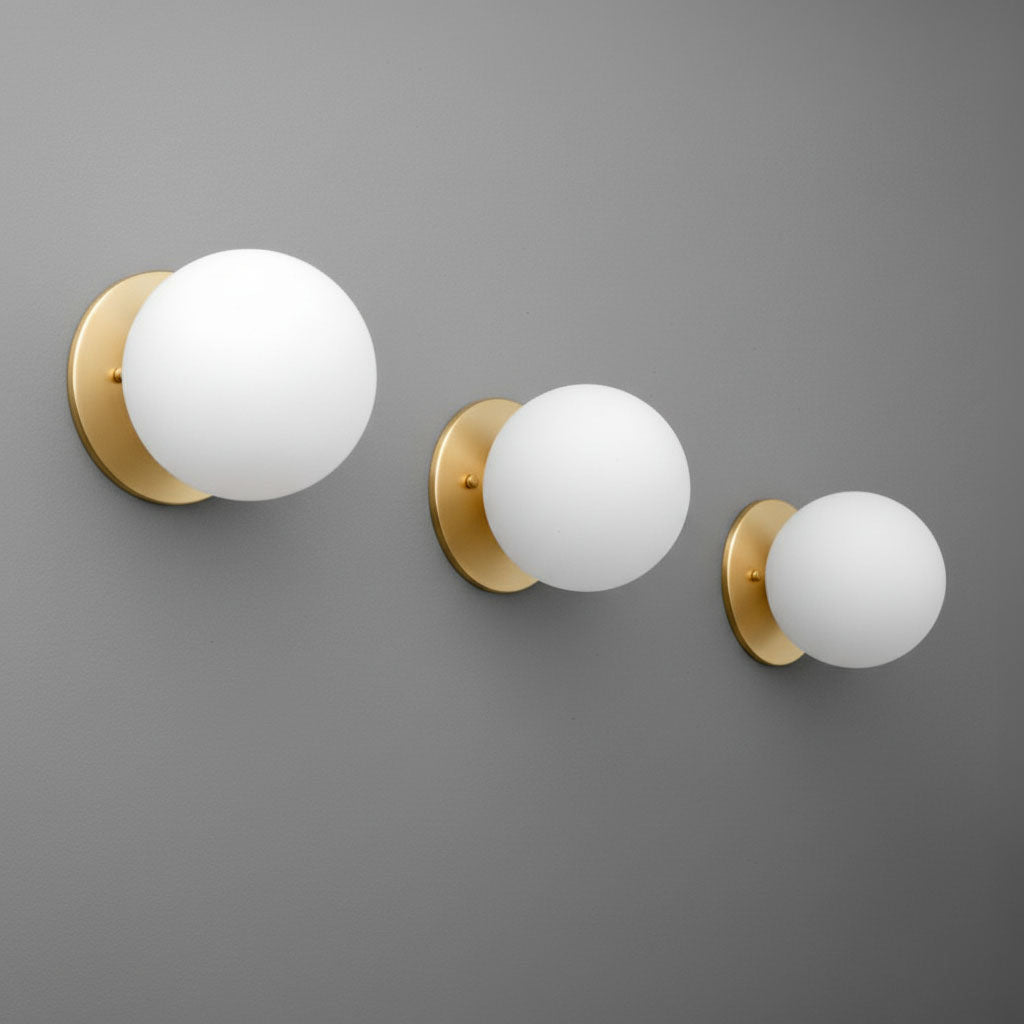 Three antique brass wall sconces with matte white glass globe shades, mounted on a gray wall, in small, medium, and large. - Sconce Model No. 5206