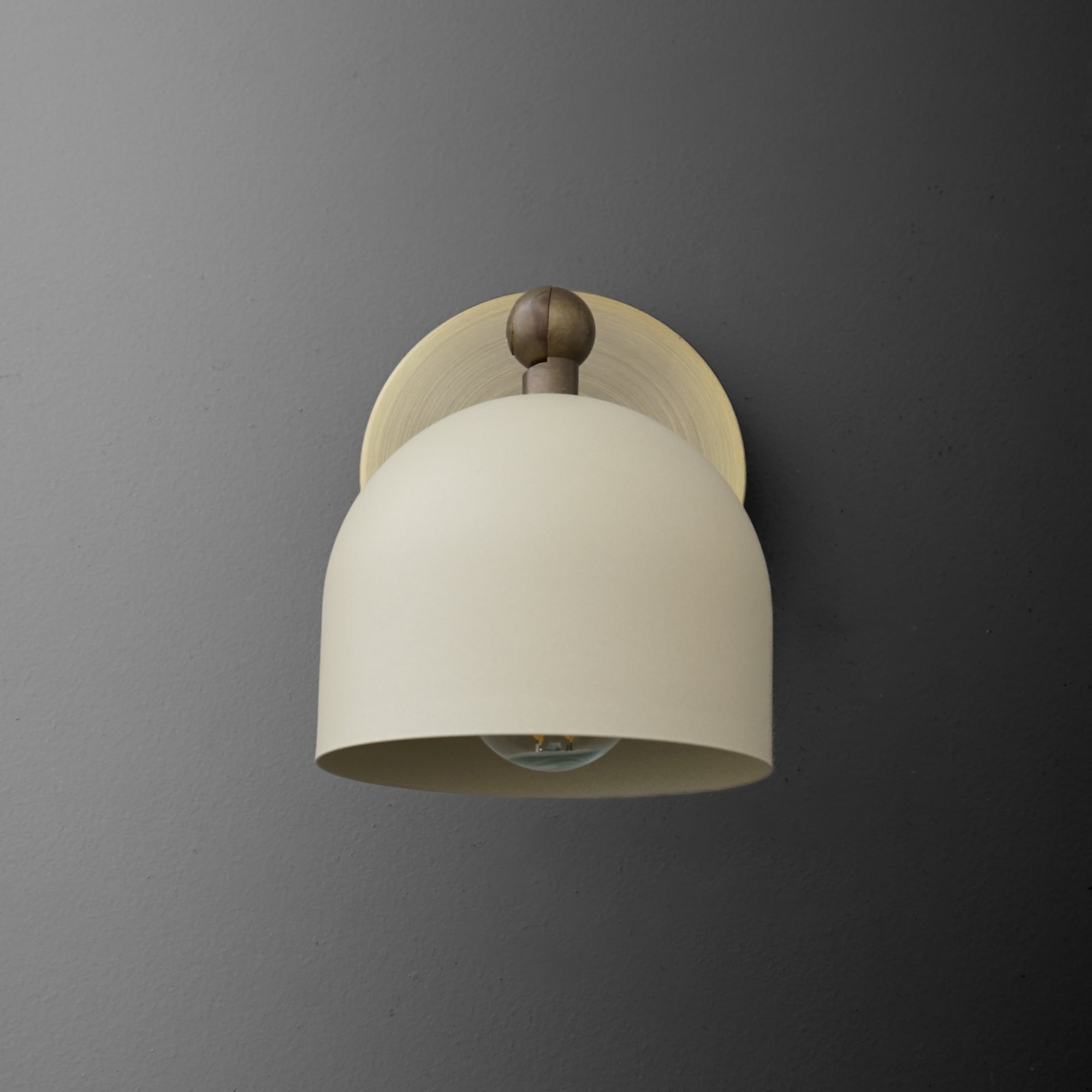 Antique brass wall sconce with cream-colored cylinder shade and round backplate on gray wall. - SCONCE MODEL NO. 5103