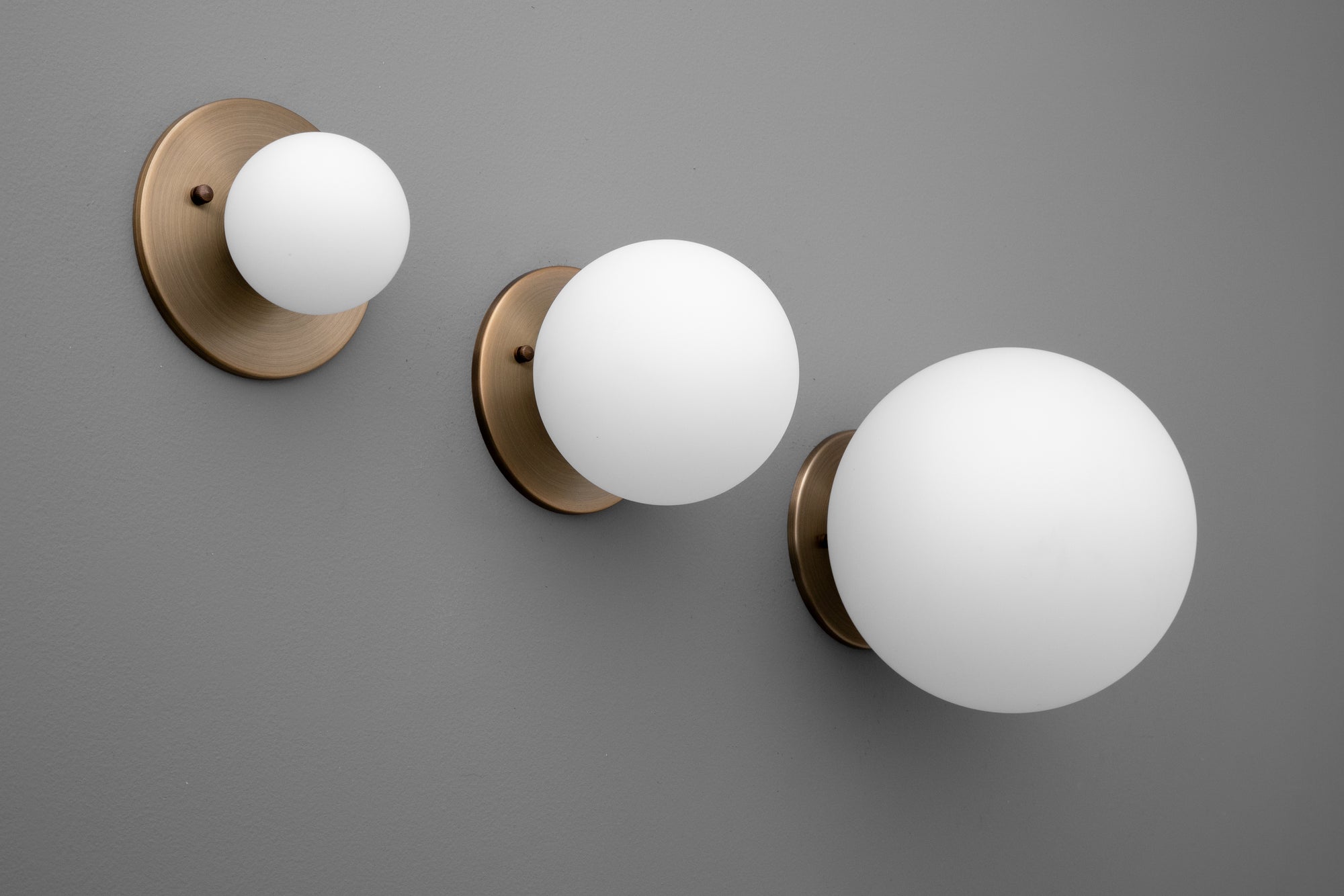 Three antique brass wall sconces with frosted white globe shades in small, medium, and large sizes on a gray wall. - Sconce Model No. 5206