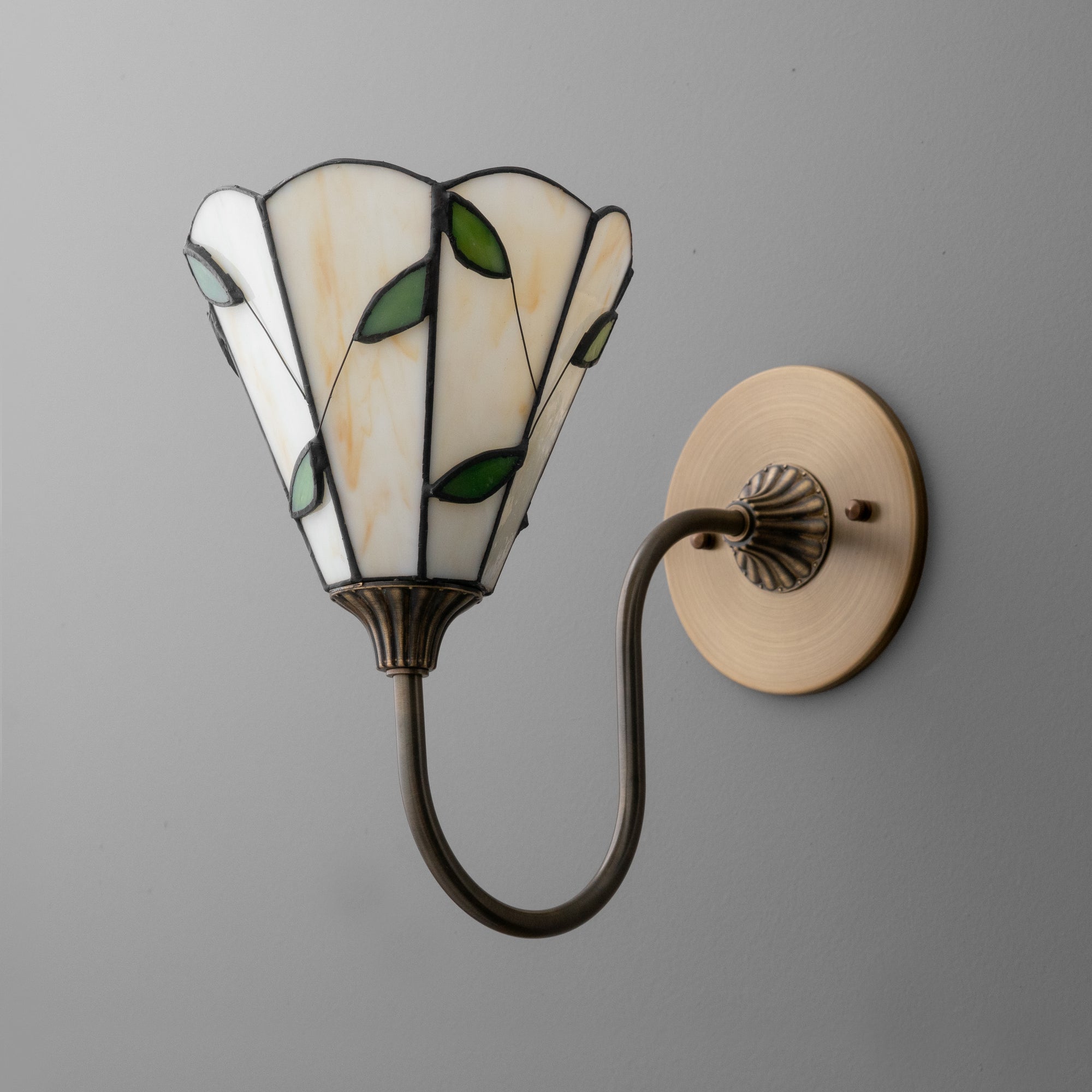 Antique brass wall sconce with curved arm and stained glass shade featuring green leaves and cream panels. - SCONCE MODEL NO. 1053