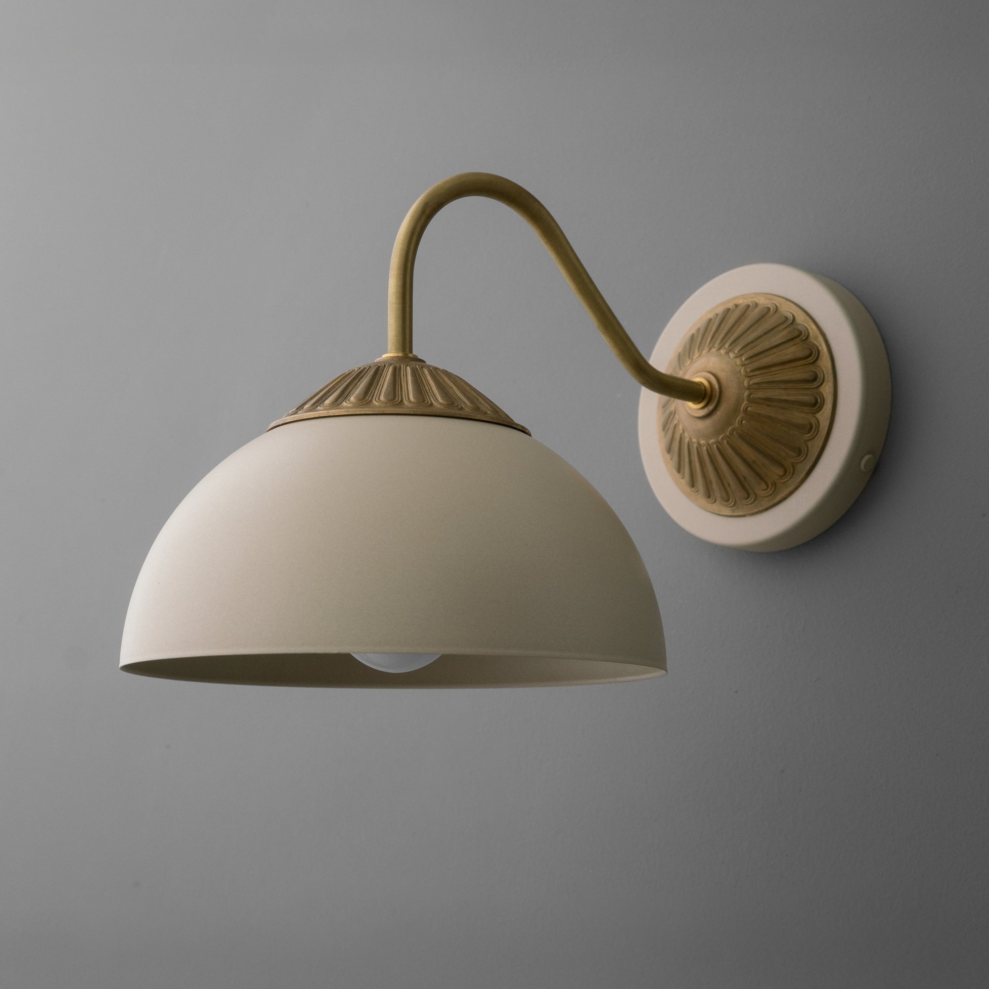 Raw brass wall sconce with curved arm and cream dome-shaped shade against gray wall. - SCONCE MODEL NO. 9408