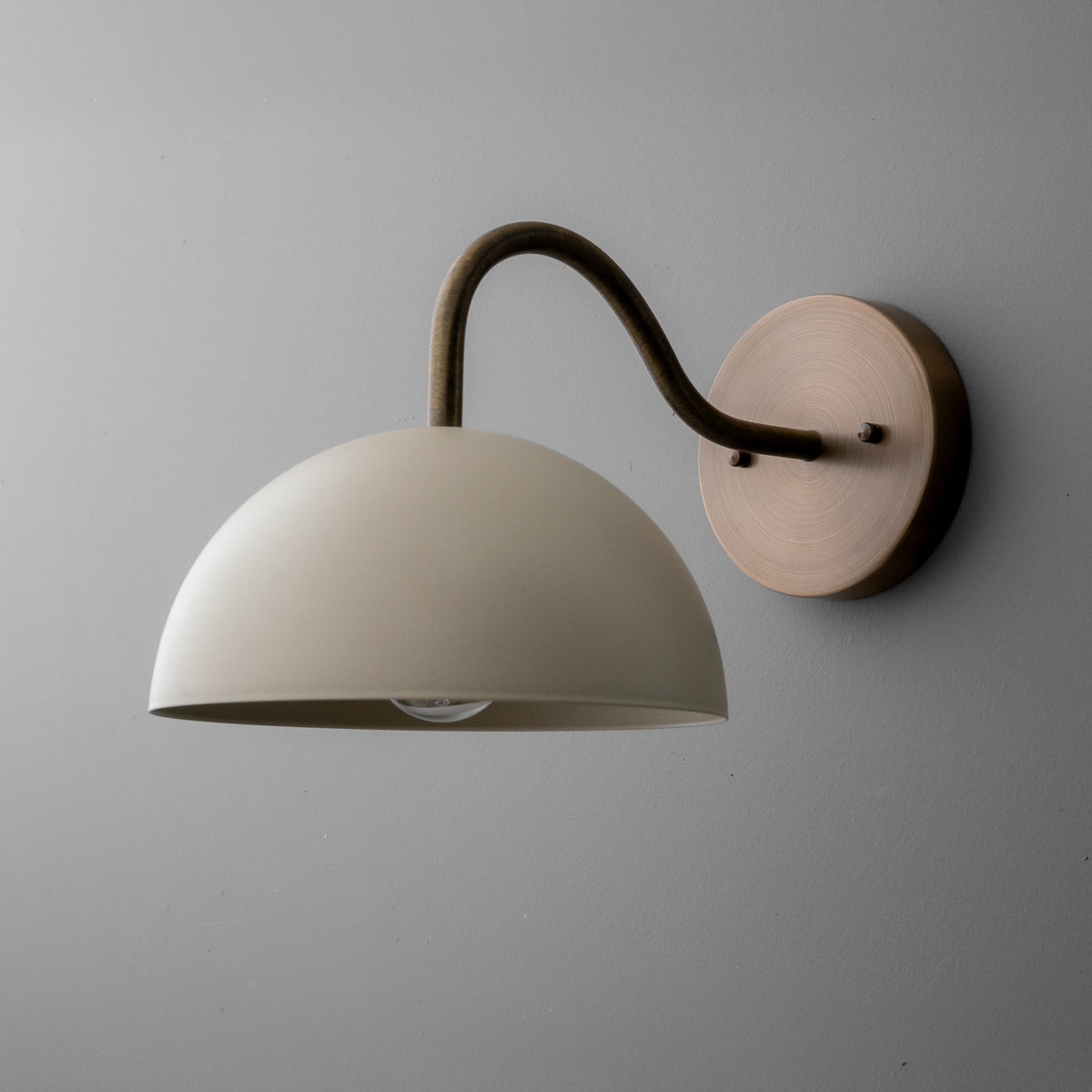 Wall sconce with a cream dome shade and curved raw brass arm mounted on a round brass base. - SCONCE MODEL NO. 6679