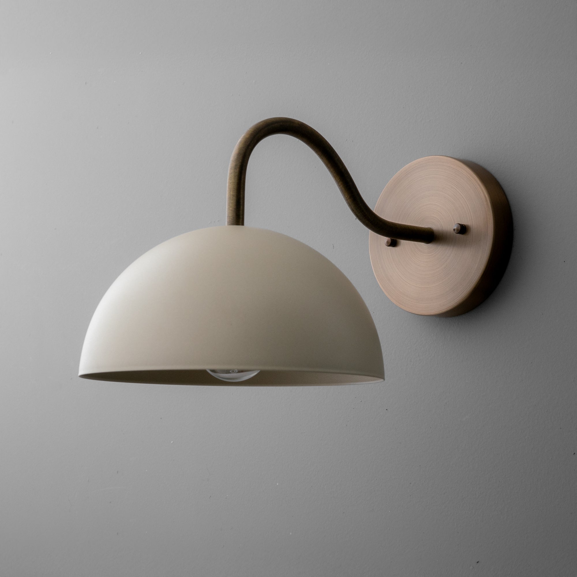 Wall sconce with a cream dome shade and curved raw brass arm mounted on a round brass base. - SCONCE MODEL NO. 6679