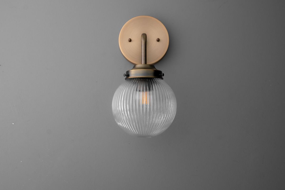 Wall sconce with ribbed glass globe and antique brass finish mounting plate against gray wall. - SCONCE MODEL NO. 6857