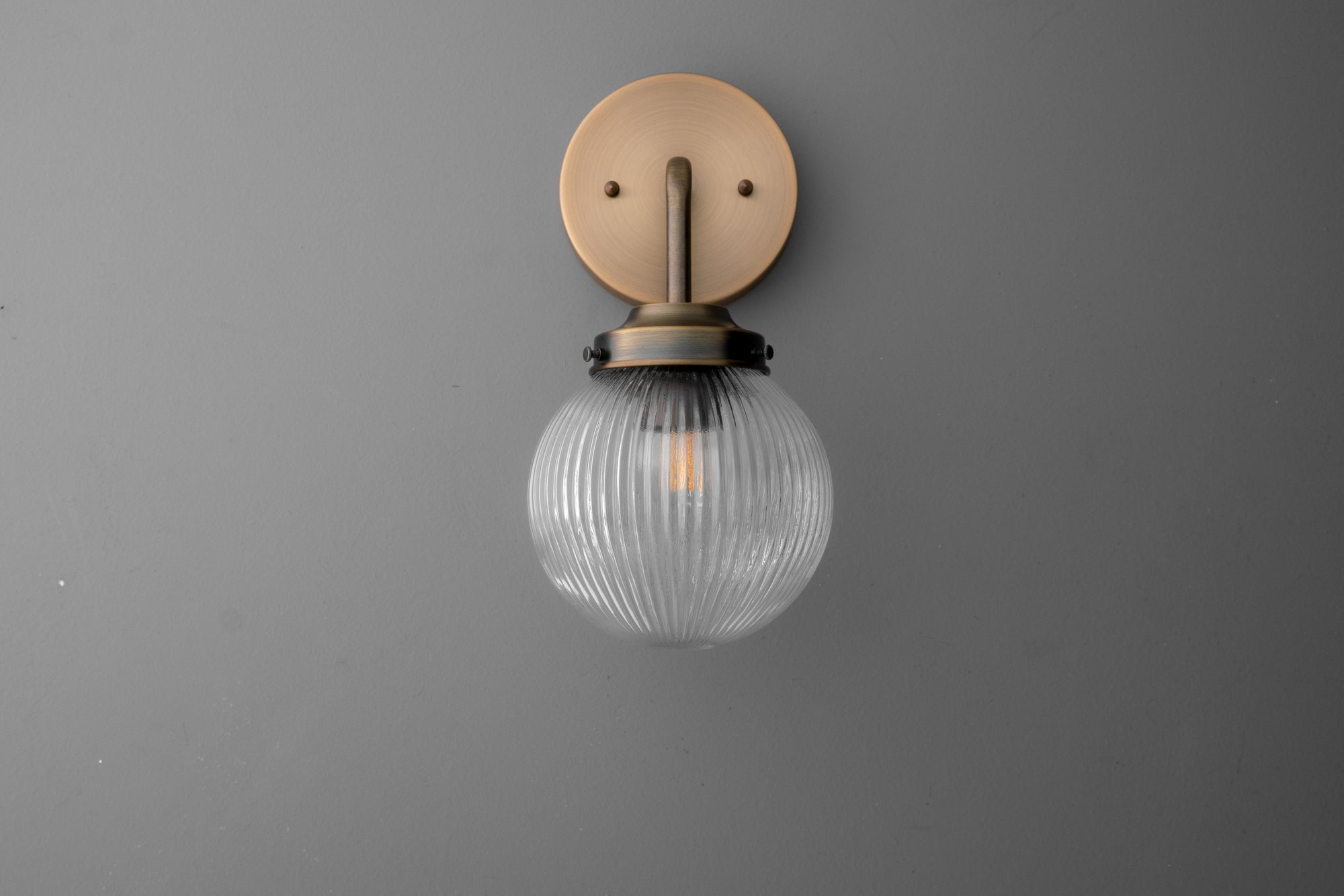 Wall sconce with ribbed glass globe and antique brass finish mounting plate against gray wall. - SCONCE MODEL NO. 6857