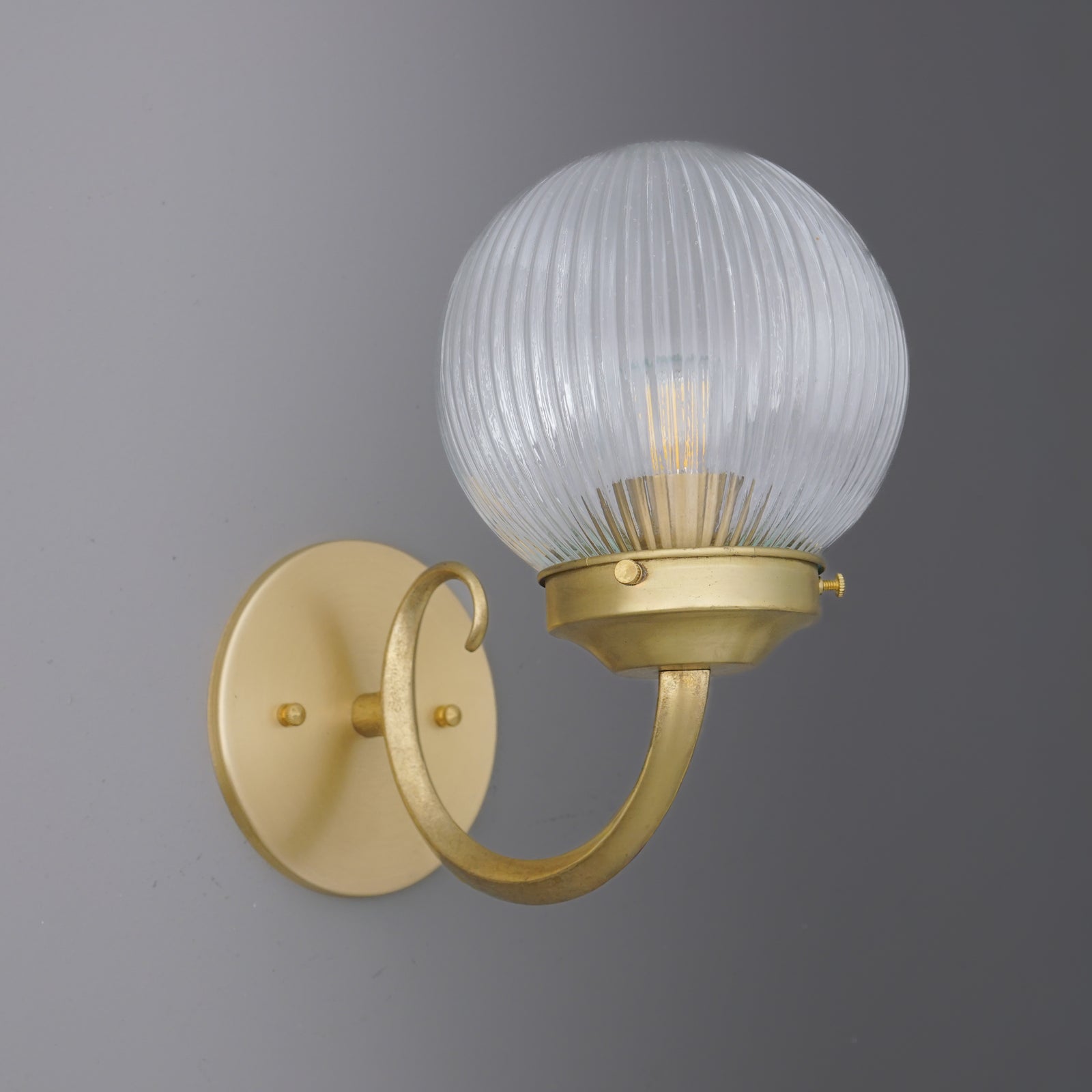 Matte gold metal wall sconce with curved arm and ribbed clear glass globe shade with visible bulb. - SCONCE MODEL NO. 1093