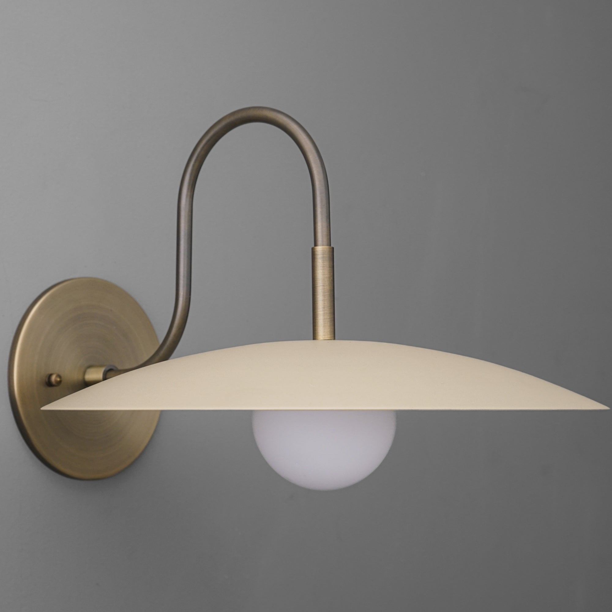 Antique brass wall sconce with curved arm and cream flat dome shade over frosted globe bulb. - SCONCE MODEL NO. 9171