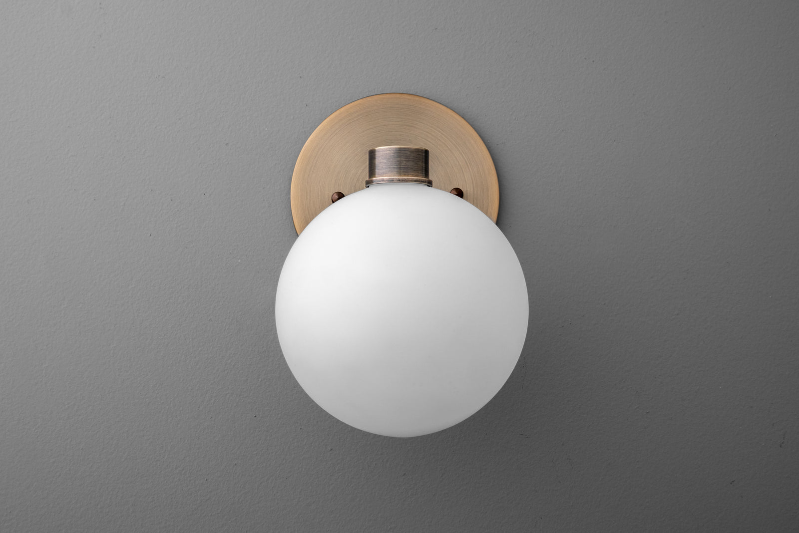 Wall sconce with frosted white glass globe and antique brass round backplate on gray wall. - SCONCE MODEL NO. 2341