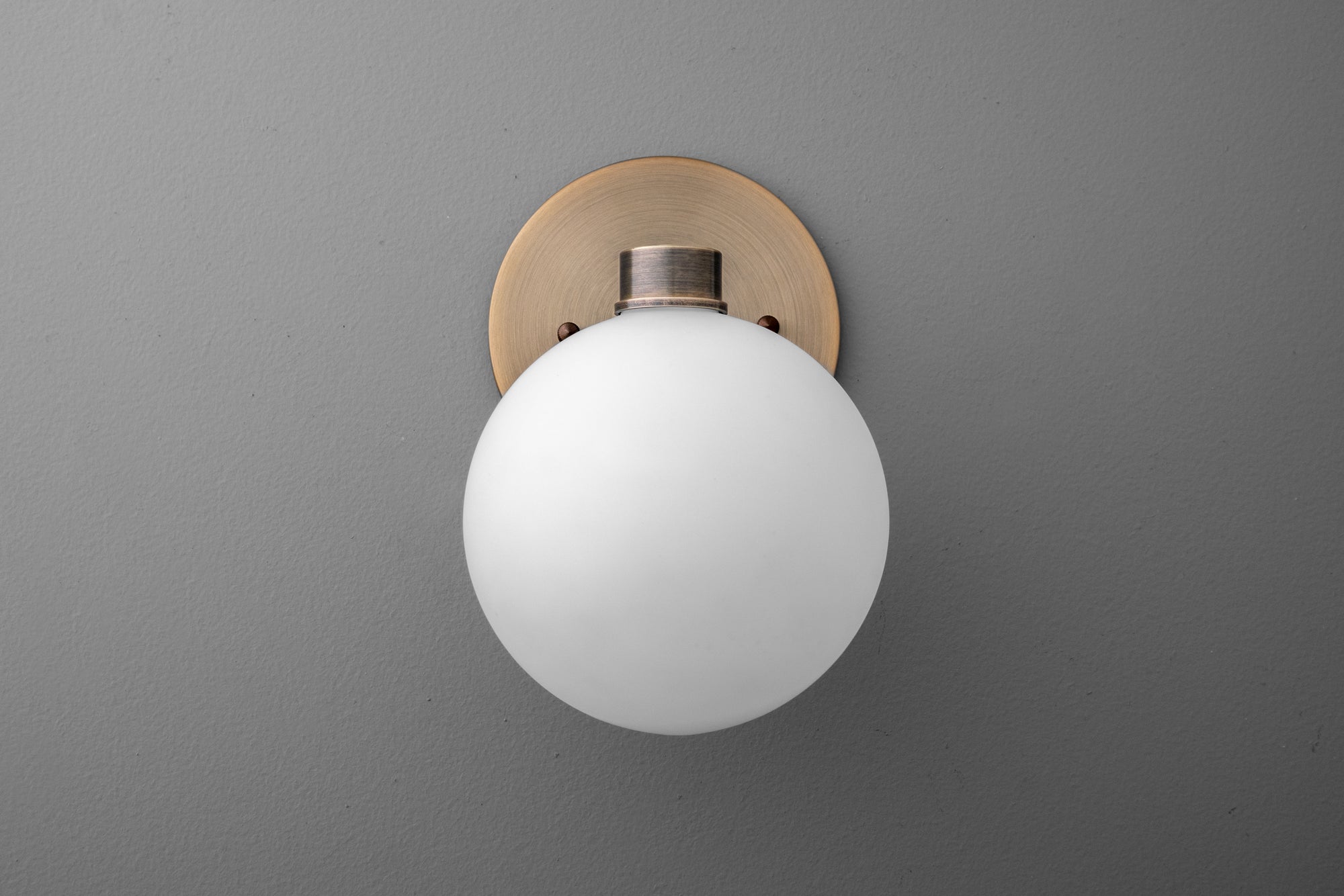 Wall sconce with frosted white glass globe and antique brass round backplate on gray wall. - SCONCE MODEL NO. 2341