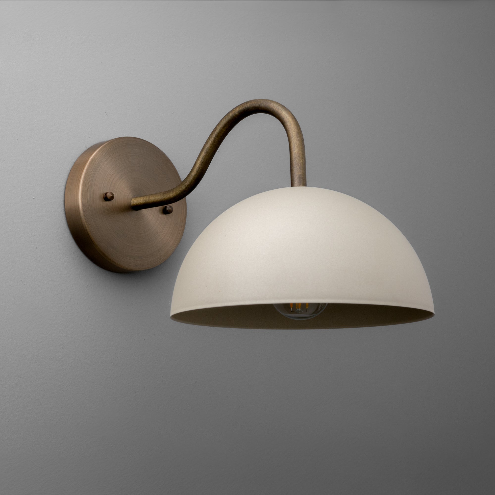 Wall sconce with raw brass curved arm and round cream dome shade mounted on gray wall. - SCONCE MODEL NO. 6679